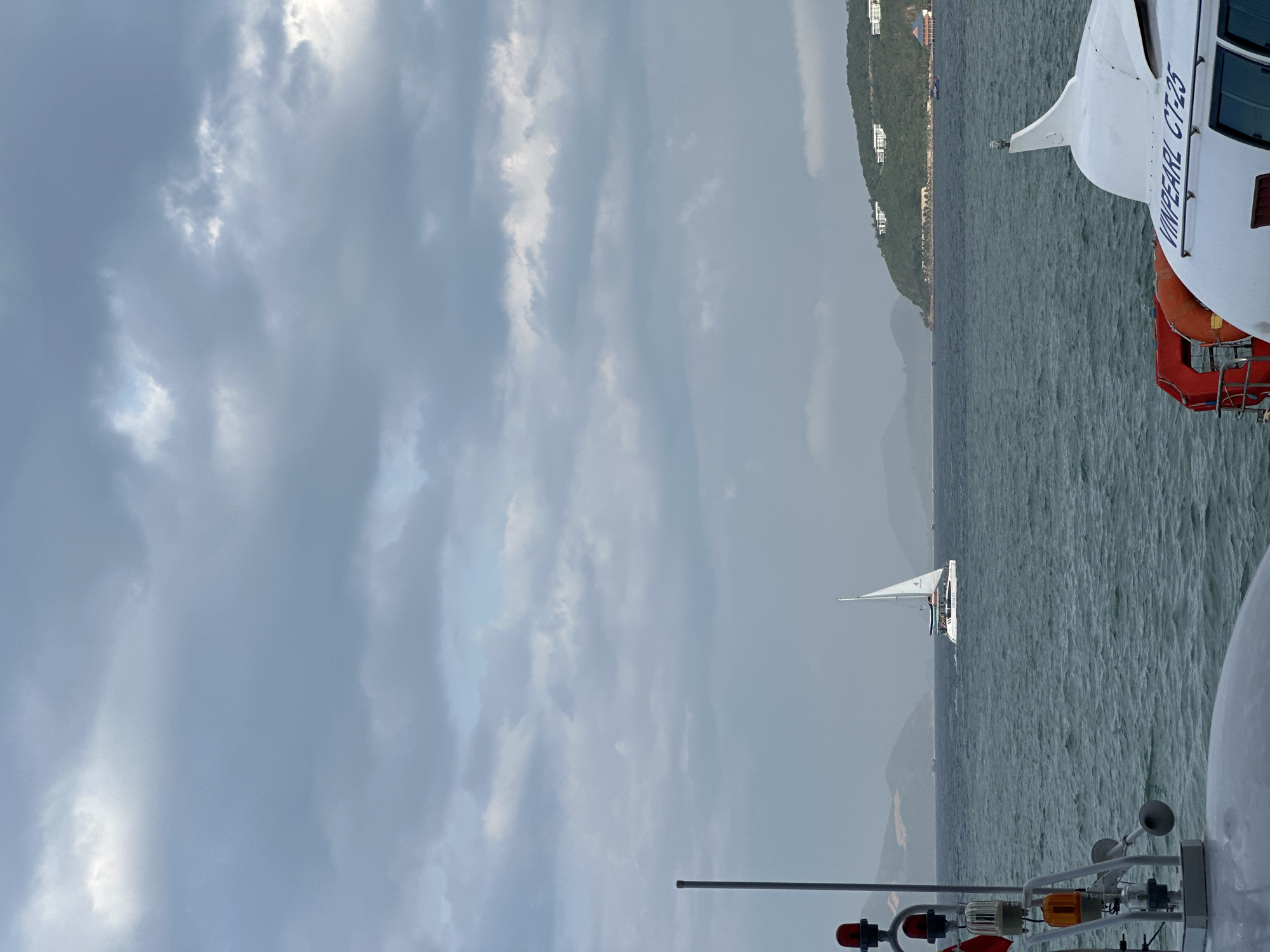 The image displays a maritime scene in the coastal waters of Nha Trang Ward, Vietnam, viewed from the deck of a vessel.
In the immediate foreground on the left, a grey vertical pole supports a stack of two red lights and one amber light, fixed to a white mounting bracket. On the right foreground, the white hull and upper structure of a boat are visible, featuring the blue inscription "VINPEARL CT-25." A red life raft holder or rack is attached to the vessel, containing a circular orange life buoy.
The middle ground consists of choppy, blue-green water. A white catamaran sailboat, equipped with a single mast and a white triangular sail, traverses the water from left to right. Figures are faintly discernible on board the sailboat.
The background reveals distant landmasses. To the far left, hazy, blue-grey mountains or hills recede into the distance. To the right, a green, forested island or peninsula is visible, dotted with several white buildings, likely resorts or residences, on its slopes and near the shore; a distinct building with a red roof is also present at the water's edge.
The sky is predominantly overcast with varying shades of grey clouds, and lighter patches of blue are visible where the cloud cover is thinner.
