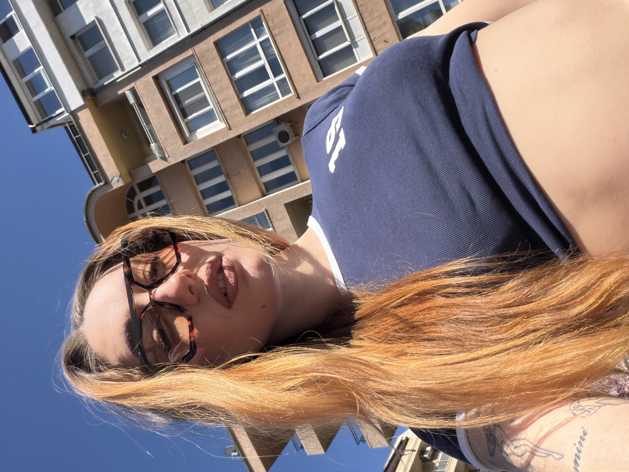 A low-angle, upward-facing shot depicts a fair-skinned woman with long, reddish-blonde hair, wearing tortoiseshell glasses. She is positioned in front of a building with multiple windows, suggesting an urban setting. The building appears to be a multi-story structure with light-colored walls and windows, typical of architecture in Sofia, Bulgaria. Her gaze is directed toward the camera, creating a sense of direct engagement. She is wearing a dark blue top. Part of a tattoo is visible on her arm. The clear blue sky is visible in the background, suggesting a sunny day.