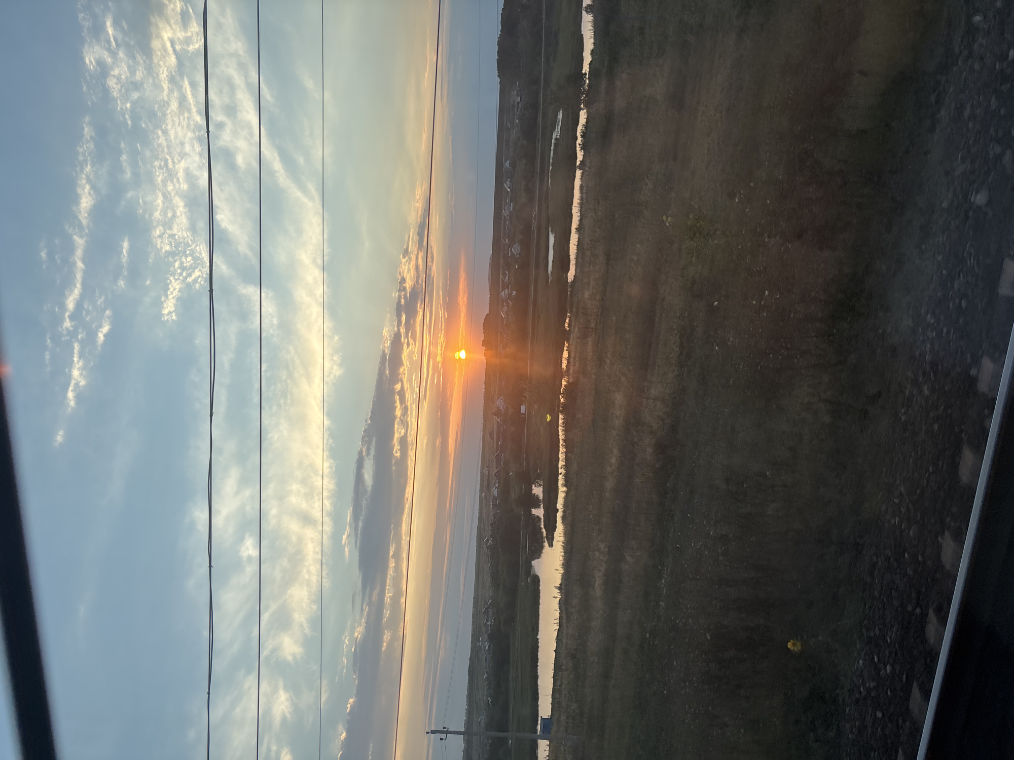 The image depicts a rural landscape at sunset, with a view of scattered houses and a body of water. The sky is a blend of blue and orange, with wispy clouds. Several utility wires run across the frame, creating horizontal lines. The foreground is dominated by a grassy field, and in the distance, a small cluster of buildings is visible. There are no people or animals in the image. The scene appears to be set in a quiet, undeveloped area. The city of Abdulino, Russia, is known for its agricultural landscapes and rural settlements, consistent with the scenery presented in the photograph.