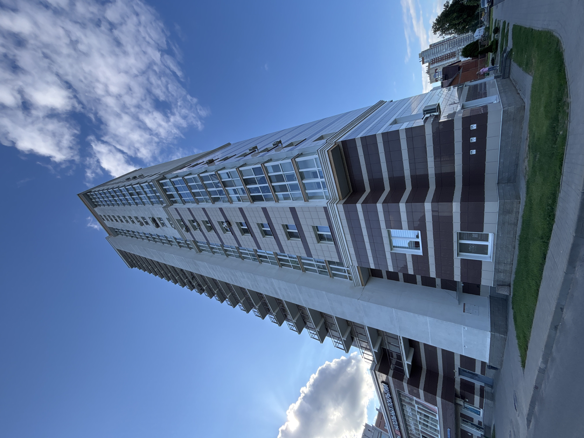 The image displays a tall, modern apartment building in Minsk, Belarus. The building has a distinctive facade with alternating horizontal bands of brown and beige. Numerous windows and balconies are visible along its length. The sky is a vibrant blue with scattered white clouds, suggesting fair weather. The sun is shining brightly, casting a slight glare on the building's reflective surfaces.
The scene appears to be a residential area, with a paved walkway and a well-maintained lawn bordering the building. In the background, other buildings, including some taller, more slender structures, are visible. One of these buildings has a reddish-brown brick facade. A few trees are also present.
On the ground level of the apartment building, there appears to be commercial space, indicated by windows and signage. One piece of visible text on the building in the lower right corner, on a sign with red lettering, appears to be in Russian or Belarusian, and partially reads "НАПОЛЬНЫХ ПОКРЫТИЯ" which translates to "floor coverings." Another sign above a window on the same level reads "PRO STUDIO" in English. There are two people visible on the sidewalk near the entrance of the building, one wearing a pink shirt and the other a blue shirt.
The image is taken from a low angle, emphasizing the height of the apartment building and looking up towards the sky. The time of day is likely afternoon, given the bright sunlight and the angle of the shadows. The overall impression is one of a well-developed urban environment on a pleasant day.