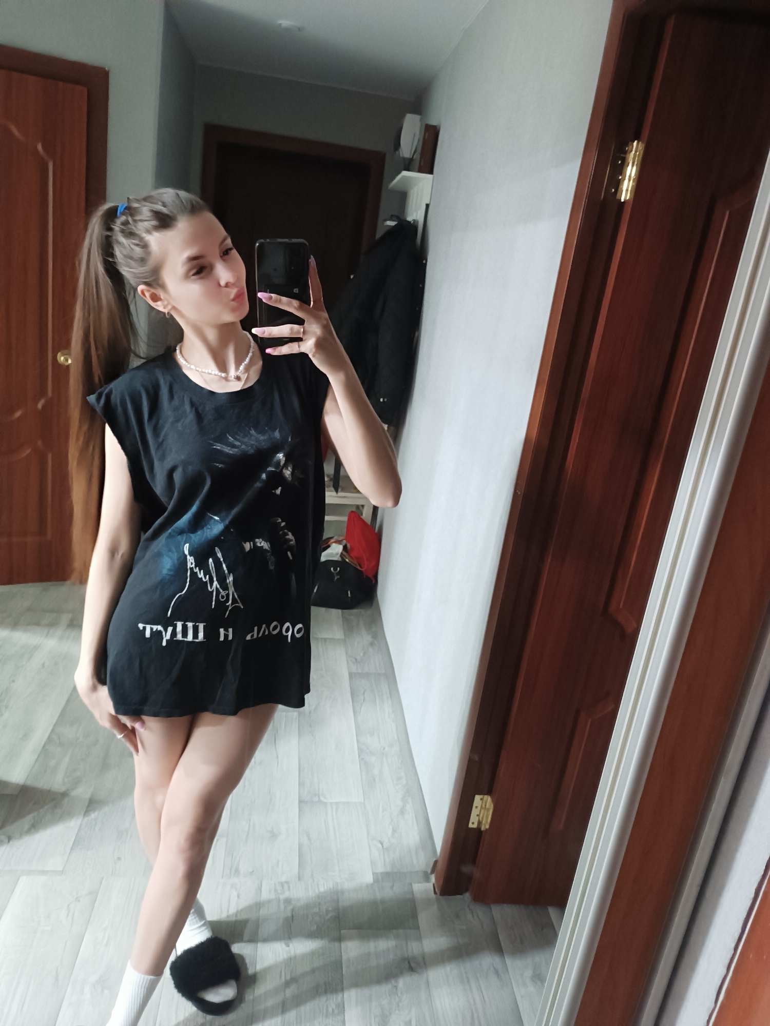 A young woman is posing for a selfie in an indoor environment. She has long brown hair pulled back in a ponytail with a blue scrunchie. She wears a black oversized graphic t-shirt, a pearl necklace, white socks, and black fuzzy slippers. She is holding a smartphone up to take the photo and making a kissy face.
The background includes a wooden door on the left, a gray wall with a shelf and some items on it, a coat hanging, and another wooden door on the right reflecting the scene. The floor is light gray with a wood-like pattern.
The location is suggested to be in Chaikovsky, Russia.
