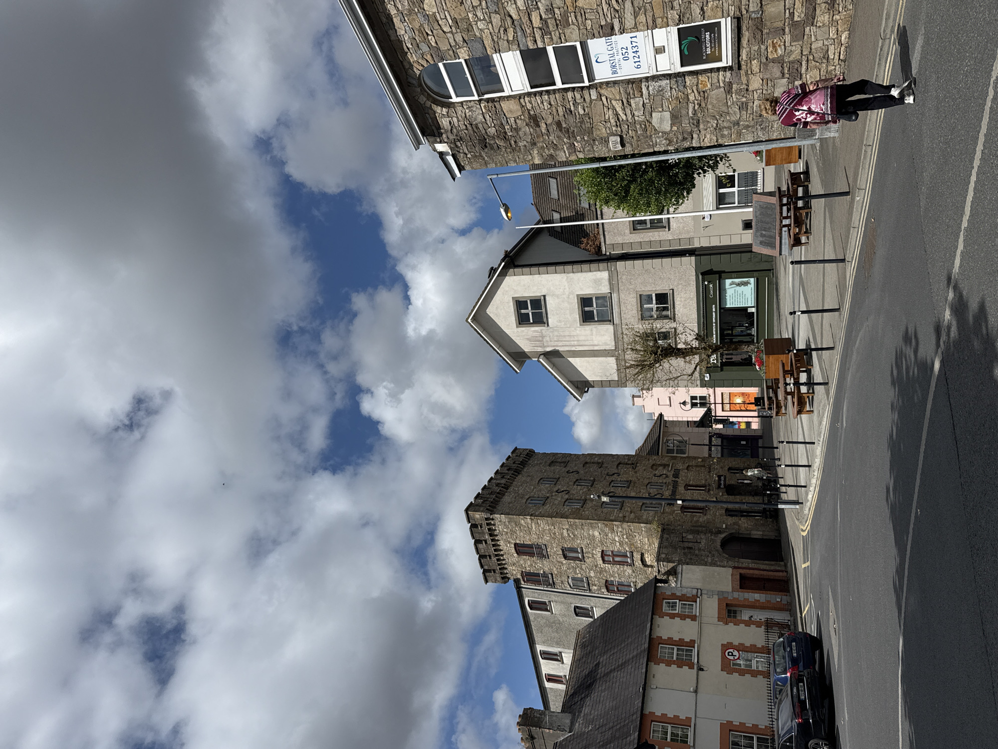 This is a street-level, eye-level, outdoor photograph taken in Clonmel, Ireland. The image captures a view of a street with several buildings and a cloudy sky. On the right side of the image, a stone building with an arched window and a sign that reads "BORSTAL GATE DENTAL PRACTICE" is visible. To the left of this building, a lighter-colored building with several windows is present. Further left, a tall, narrow stone building with a crenelated top stands prominently. Adjacent to it, a lighter-colored building with multiple windows is also visible. In the foreground, the street is paved, and there are some parked cars on the left side of the image. A woman in a patterned shirt and dark pants is walking across the street towards the right side of the frame. Several wooden picnic tables are set up on the sidewalk area in front of the buildings. The sky is filled with a mix of white and gray clouds.