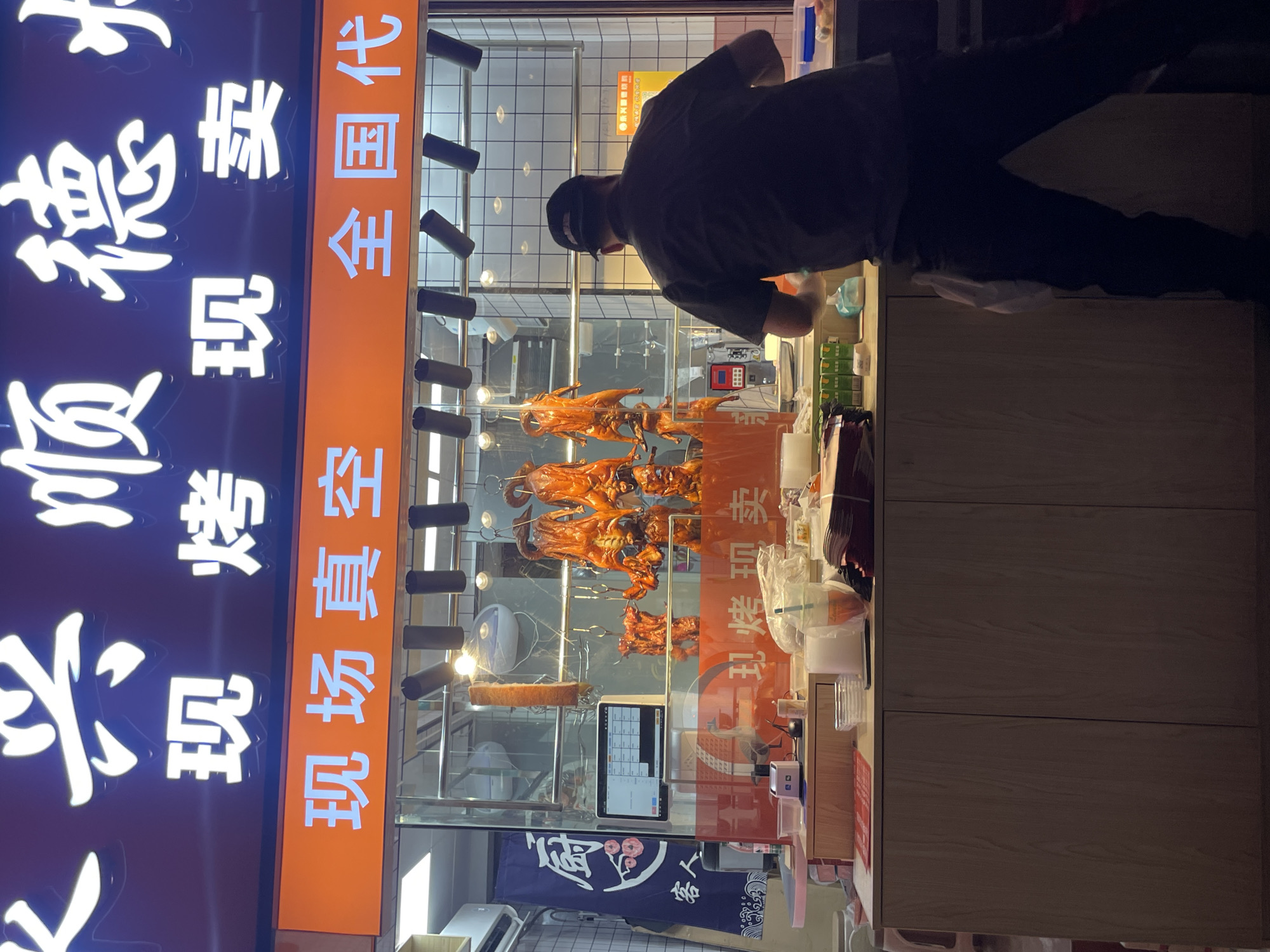 Captured in the Tianhe District of China, the scene showcases a food stall. A man, partially visible, stands behind the stall, presumably preparing food. Ducks hang inside a glass display case, possibly roasted or grilled, suggesting the stall specializes in poultry. The stall is adorned with signs featuring Chinese characters. One sign, illuminated with neon lighting, displays the name of the stall. An orange vertical sign translates to "on-site vacuum sealing, nation-wide agent". A cash register, various containers, and packaging materials are arranged on the countertop, further indicating the stall's commercial function.