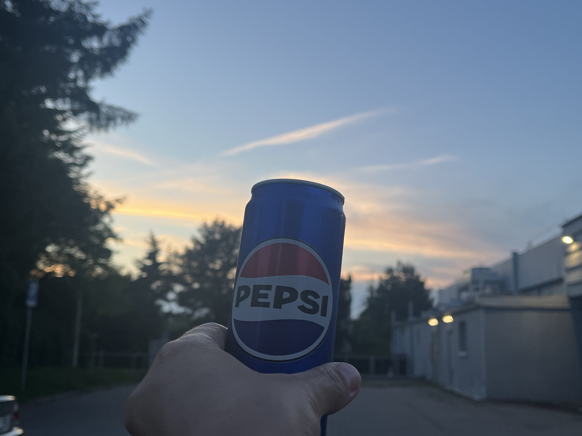 A hand is holding up a blue Pepsi can in front of a sunset sky with wispy clouds. The can has the Pepsi logo, which is a red, white, and blue circle with the word "PEPSI" written in white. In the background, there are trees and buildings, and a street or road. The scene appears to be outdoors, possibly in a suburban or industrial area, at dusk.