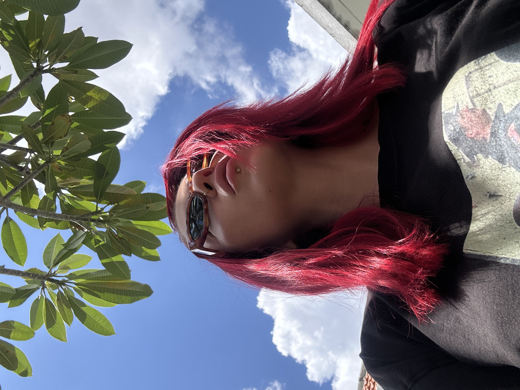 A photograph taken in North Kuta, Indonesia, depicts a woman with dyed red hair wearing sunglasses and a black t-shirt with a graphic print. She is positioned beneath a tree with large green leaves against a backdrop of a blue sky with scattered white clouds. The camera angle is tilted upwards, capturing her from a low perspective. The sky is visible between the leaves of the tree. A white surface, potentially a building element, is visible in the upper right corner. The woman has a piercing on her upper lip. The photograph is taken outdoors during daylight.