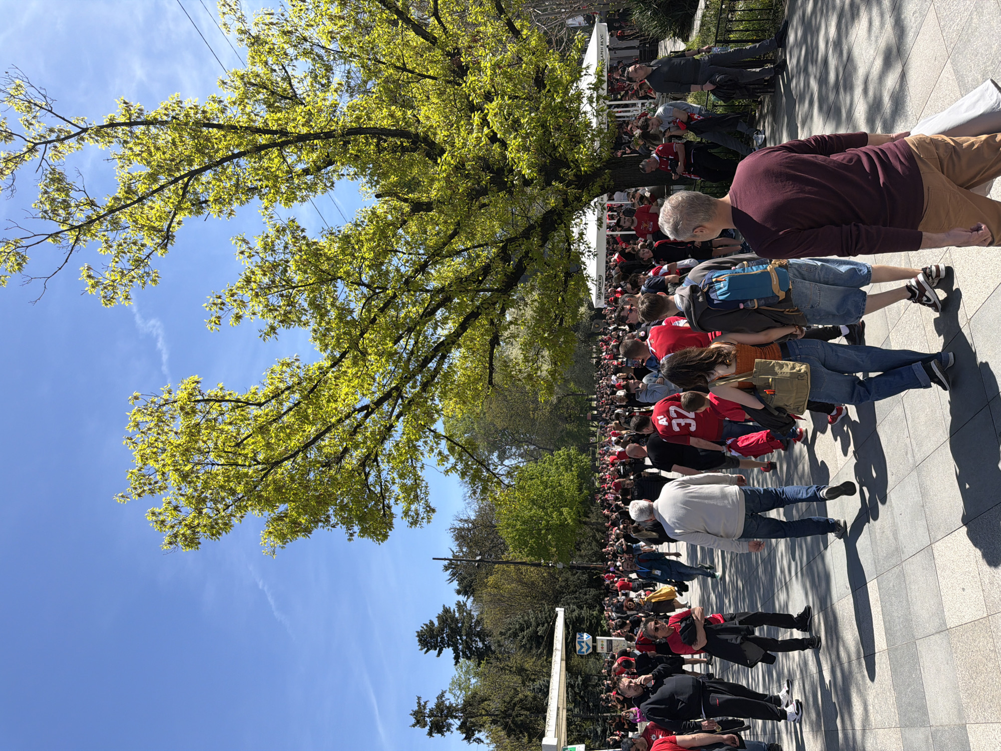 A large crowd of people lines a sidewalk in Sofia, Bulgaria, under a clear blue sky with a large deciduous tree providing partial shade. The majority of the crowd wears red, suggesting they are fans attending an event or gathering. The people are of varying ages and ethnicities. The background features other trees and structures, including white tents. The sidewalk is paved with a patterned stone surface. A subway sign is visible in the middle. The scene appears to be a public space filled with people.