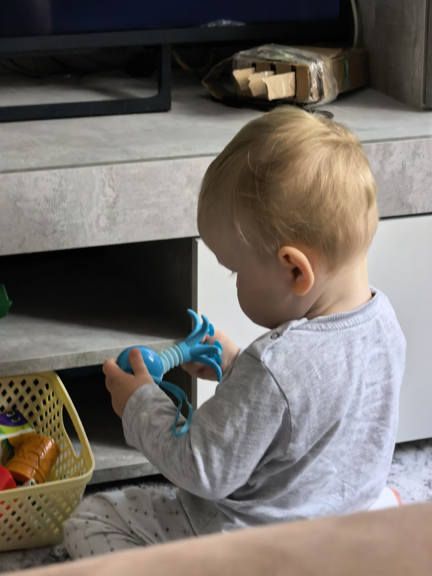 A toddler with blonde hair wearing a grey long-sleeved shirt and patterned pants is playing with a blue toy in front of a grey and white shelf. A yellow basket containing various toys is on the shelf next to the child. In the background, a television and a partially visible cardboard box are present. The image appears to be taken indoors. There is no verifiable information to suggest a specific location like Sterlitamak, Russia.