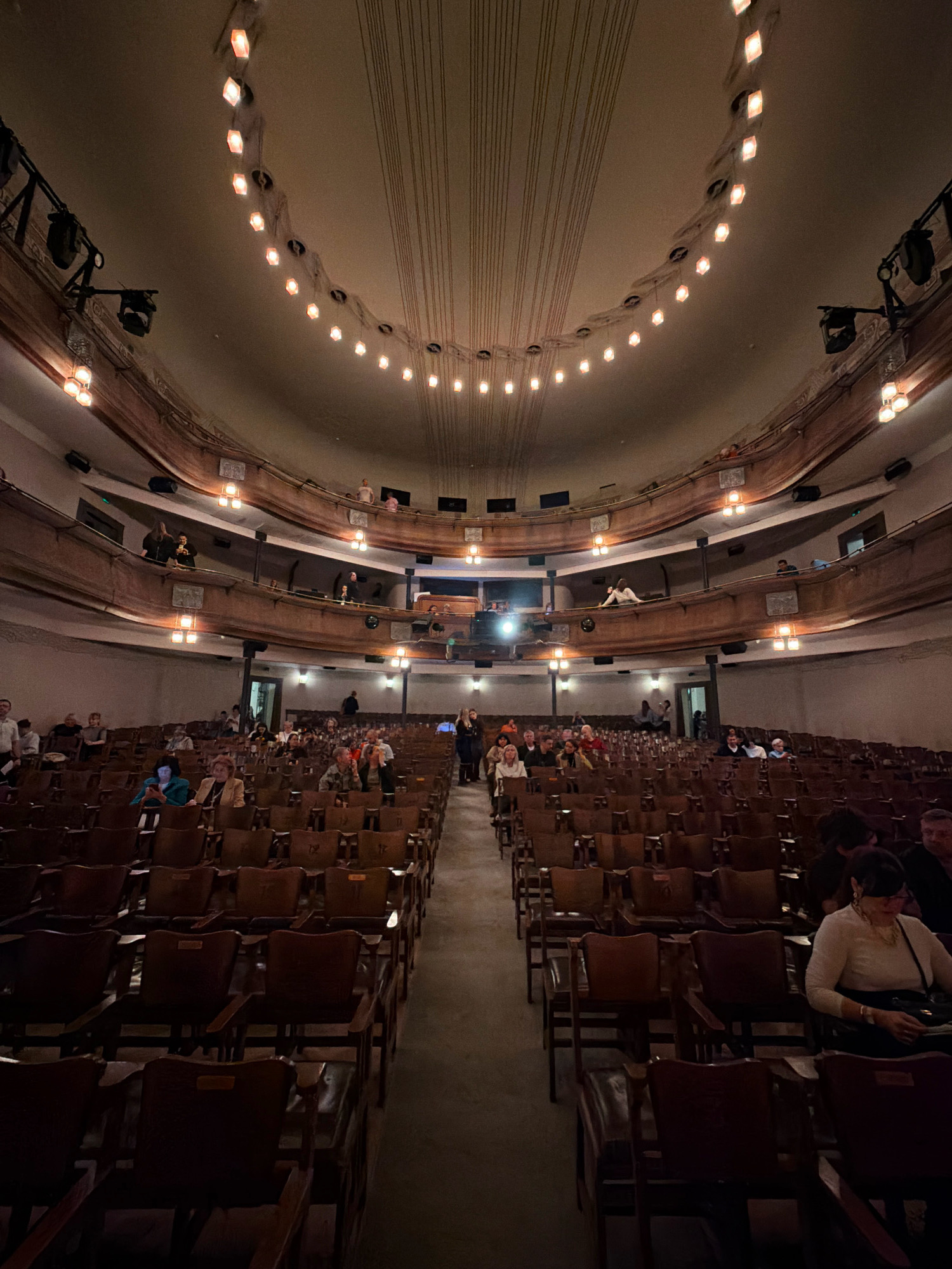 The image depicts the interior of an old theater, possibly in Moscow, Russia, judging by the architectural style and ornate detailing. The scene is illuminated by warm, ambient lighting from decorative fixtures along the curved ceiling and balconies, casting long shadows. The seating area is filled with rows of dark wooden chairs, some of which are occupied by an audience dressed in casual attire. The audience members appear to be seated, some looking forward, others engaged in conversation. The balconies above also show individuals present, observing the performance or gathering area. The overall atmosphere suggests a cultural event or performance is underway or about to commence.