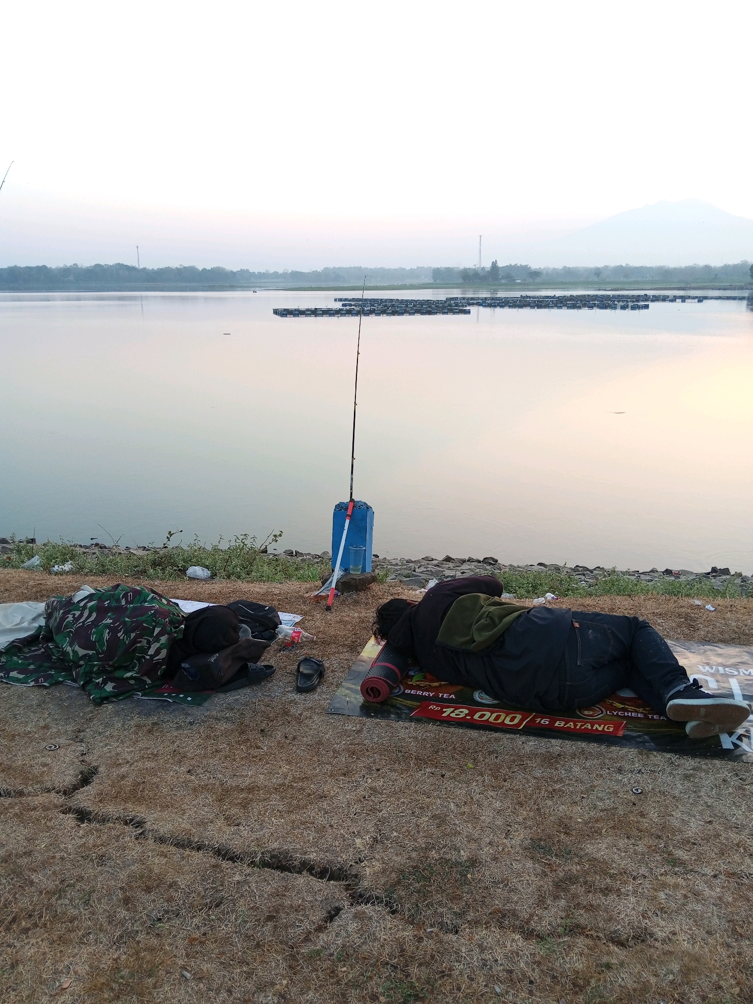 The image depicts a waterscape with a tranquil lake or reservoir in the background, likely at dawn or dusk due to the soft, diffused light. In the foreground, two individuals appear to be sleeping outdoors on a dry, grassy bank beside the water. To the left, one person is covered with a camouflage blanket, with various bags and a pair of flip-flops nearby. A fishing rod is planted upright in the ground next to a blue container filled with liquid, suggesting that fishing might be a planned activity. To the right, another person is lying on their back on a mat, dressed in dark clothing. The mat displays text and graphics, including "BERRY TEA" and a price indicating "Rp 18.000 / 16 BATANG". The environment is natural, with some vegetation and rocky terrain along the water's edge. The presence of floating structures, possibly fish farms or platforms, can be observed in the water. In the distance, a hazy treeline and a mountain are visible. The scene conveys a sense of stillness and outdoor repose.