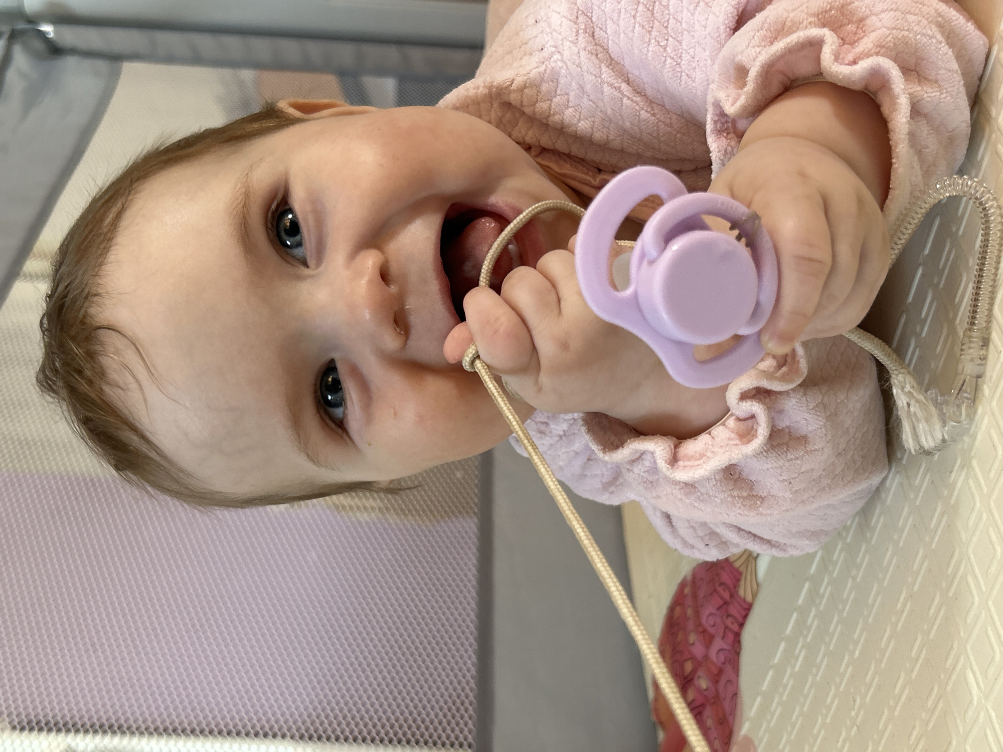 A baby wearing a pink ruffled shirt is lying down, possibly in a crib. The baby is holding a purple pacifier with a beige cord attached. The baby is looking upwards with their mouth open. The location is unknown, and there is no information in the image to suggest that it is in Rusalevka, Russia.
