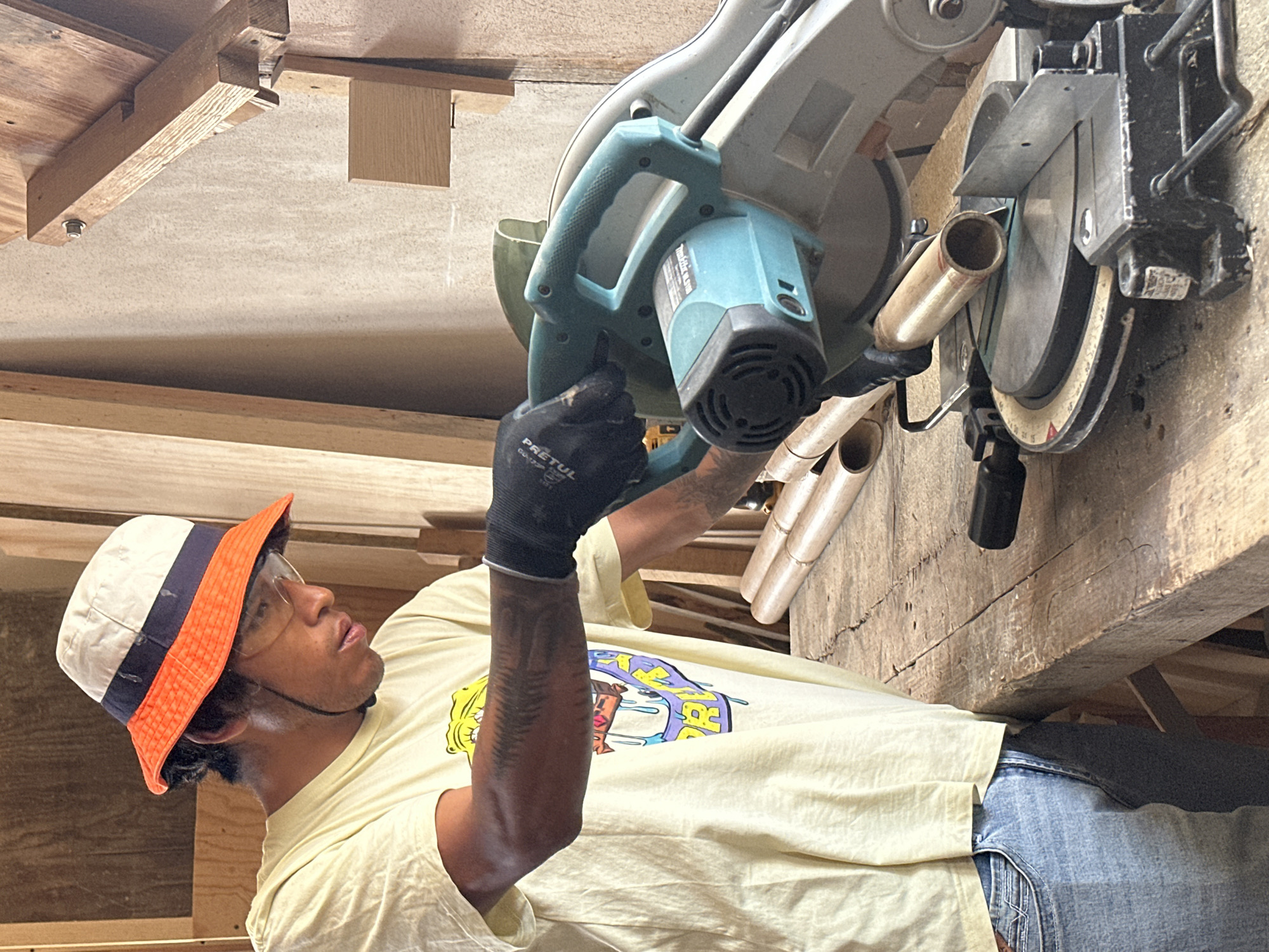 A man in a cream-colored T-shirt with a graphic design, blue jeans, a cream and orange bucket hat, and glasses is using a miter saw to cut a small, round piece of beige tubing. He is wearing a black work glove on his left hand. The miter saw is a blue and gray color. The setting is a cluttered workspace with wooden beams and construction materials overhead, possibly in a garage or workshop. The location of San Luis Tehuiloyocan, Mexico, is not discernible from the image.