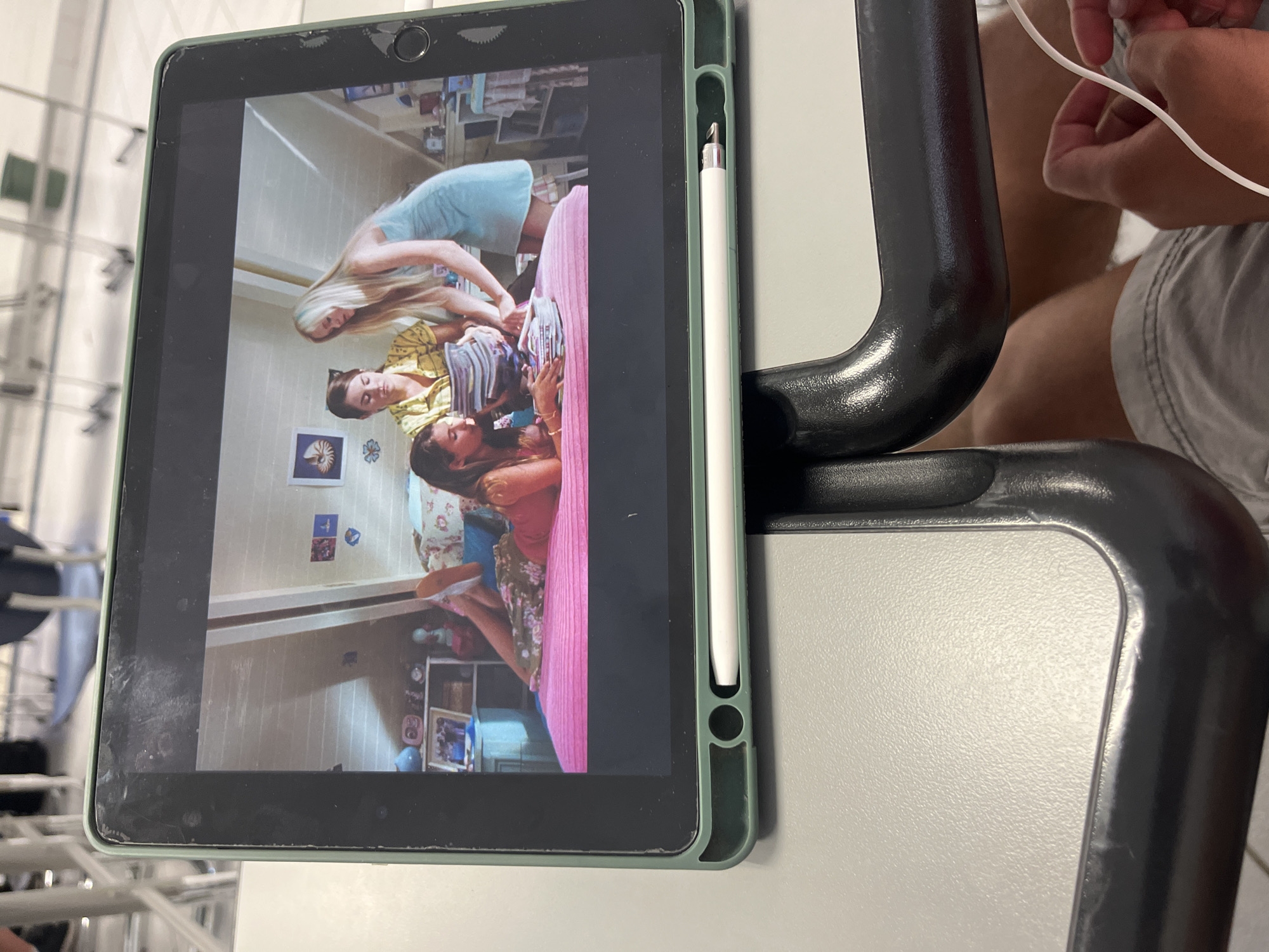 A tablet displays an image of three women in a bedroom. A white stylus is attached to the tablet's side. The tablet rests on a table next to a black metal object. A person in a gray shirt is partially visible in the background. The location is unknown.