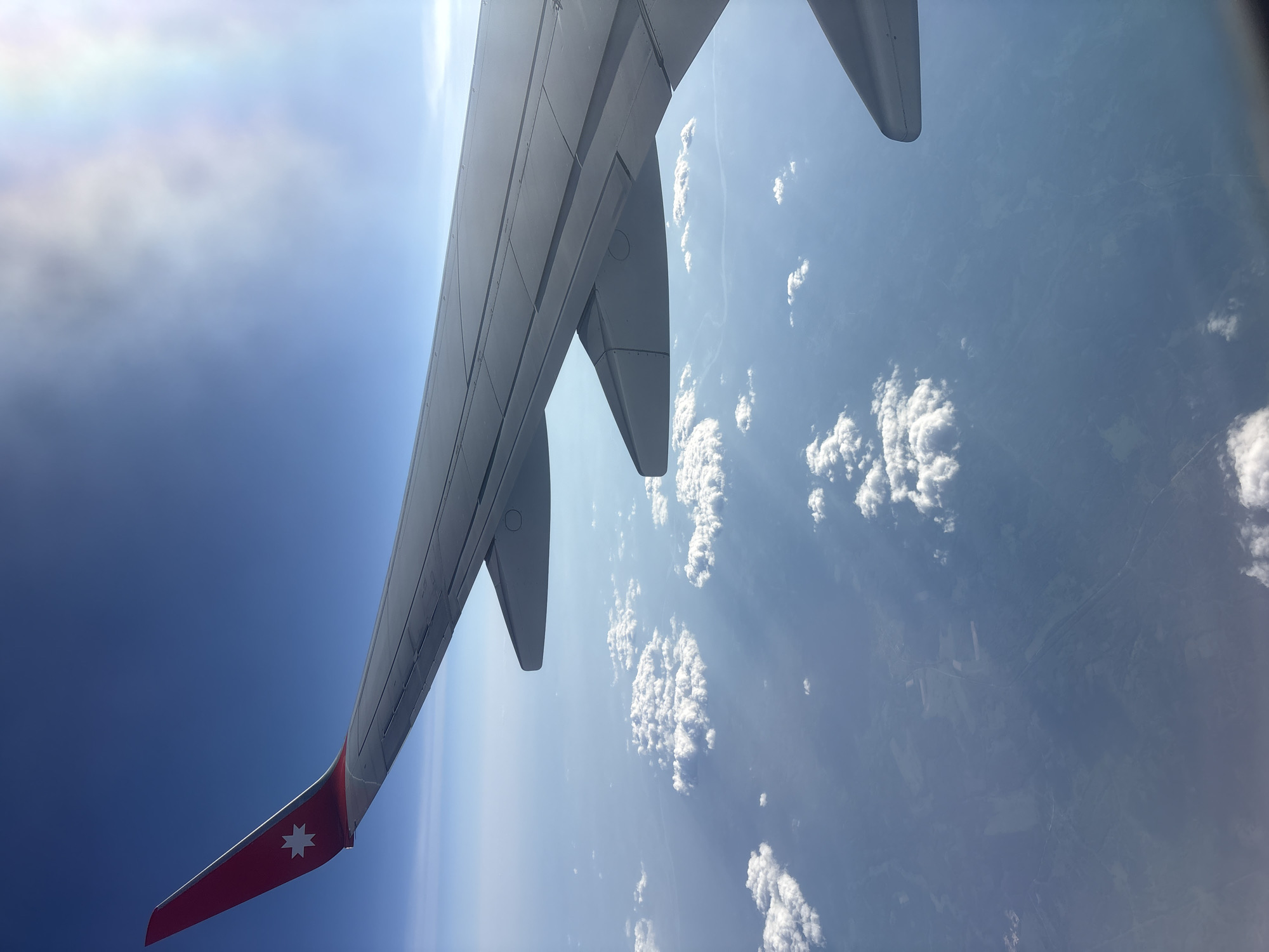The image is taken from an airplane window, showing the wing of the aircraft against a backdrop of sky and clouds. The wing is predominantly gray, with a red tip adorned with a white, eight-pointed star. The sky transitions from a deep blue to a lighter, cloudier hue, while the ground below is dotted with patches of white clouds and glimpses of land. The location is Saint Petersburg, Russia, as indicated by the star insignia on the wing.