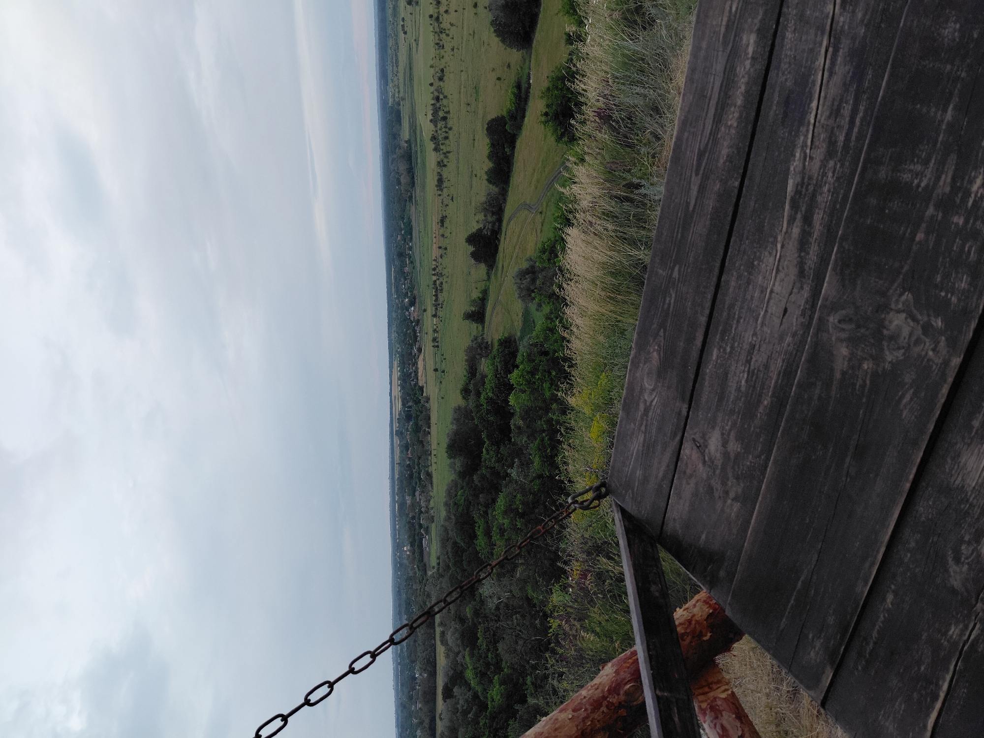 This is a high-angle, vertically oriented, full shot of a rustic wooden railing with a chain attached, overlooking a vast, green landscape under a cloudy sky. The railing, made of dark, weathered wooden planks, occupies the right side of the frame, leading the eye towards the expansive view. A thick, rusty chain is stretched across the foreground, secured to the railing and disappearing out of frame to the left.
Beyond the railing, the landscape unfolds into rolling hills covered in a mosaic of green fields and dense clusters of trees. A winding path or road is visible cutting through the fields, suggesting a rural or natural setting. In the distance, the landscape softens into a hazy horizon, with hints of more distant trees and possibly some man-made structures. The sky is overcast with soft, diffused light, indicating it could be early morning or late afternoon, or a generally cloudy day. There are no people visible in the frame. The overall mood is serene and contemplative, inviting the viewer to take in the sweeping vista. The scene appears to be an overlook or viewpoint, possibly in a park or natural reserve, offering a panoramic view of the countryside. No text is visible in the image.