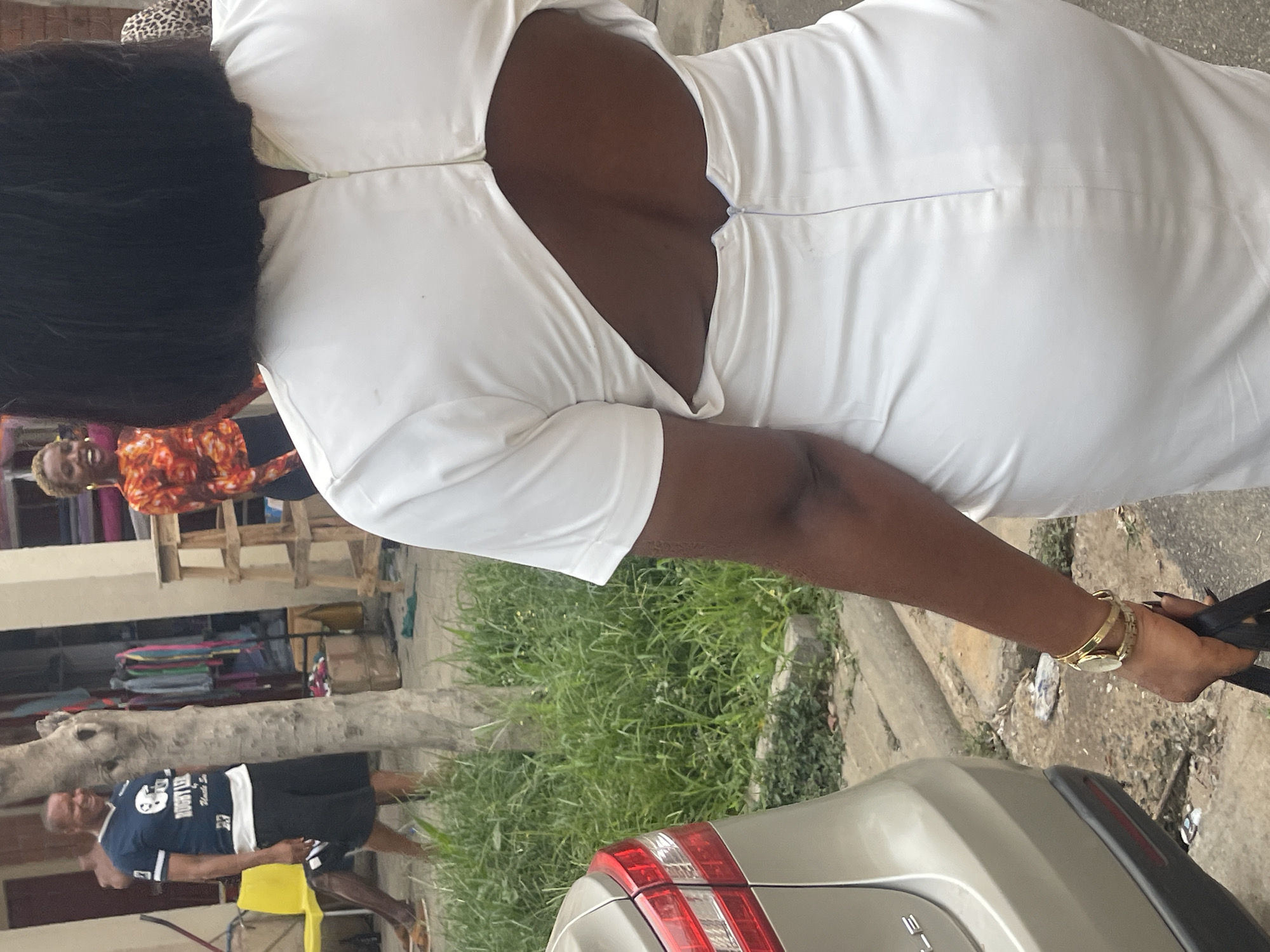 A woman with black hair wears a white dress that exposes part of her back. She has a gold watch on her left wrist and holds black objects in her hand. The rear light of a gray car is visible at the bottom right. In the background, other people are present, including one in an orange patterned shirt and another wearing a dark blue shirt with white text. A tree trunk, green foliage, and what appears to be a clothes rack are also visible in the background. The location may be in a busy area of Abuja, Nigeria, given the presence of people, a vehicle, and commercial elements like the clothes rack.