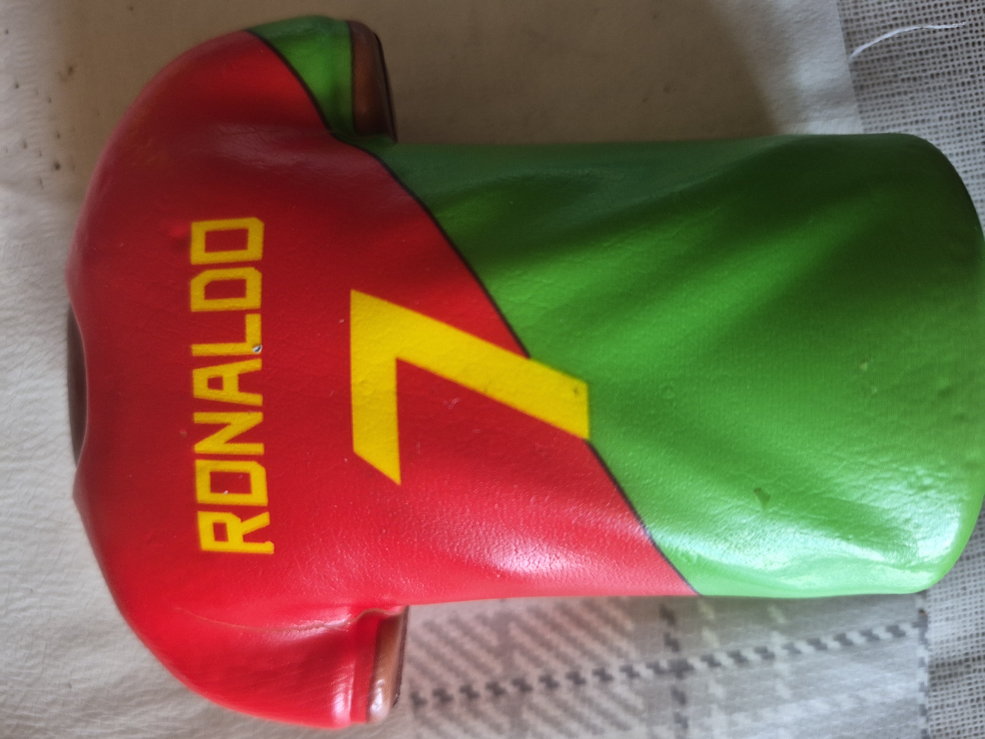 This close-up, vertical image features a decorative, miniature object resembling a football (soccer) jersey, predominantly red and green. The red section, occupying the left side of the object, prominently displays the name "RONALDO" written vertically in bold, yellow uppercase letters. To the right of the name, a large yellow number "7" is visible, indicating a tribute to the renowned Portuguese footballer Cristiano Ronaldo and his jersey number. The object transitions from red to green, representing the colors associated with the Portuguese national team.
The item appears to be crafted from a smooth, molded material, possibly plastic or ceramic, with a slightly glossy finish. The various crops reveal subtle textures and minor imperfections on its surface. The background consists of a light-colored, textured fabric, possibly a tablecloth or blanket, which shows a faint checkered or woven pattern. The lighting is even, suggesting an indoor setting. There is no specific activity taking place, as the image is a still life focusing on the object. This fan merchandise, celebrating a global sports icon, was photographed in Yerevan, Armenia.