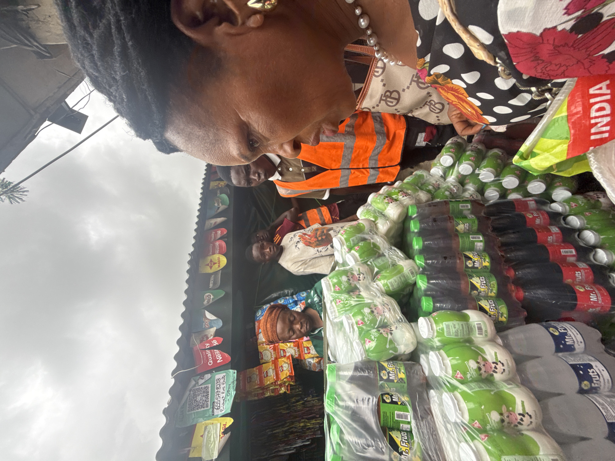 In Lagos, Nigeria, a woman wearing a pearl necklace and a patterned top stands in the foreground, looking to the right. She has dark hair pulled back and wears gold earrings. Behind her, a market stall is laden with various products, including bottled drinks and packaged snacks. Several people are visible in the background: a woman in a headscarf and green top appears to be tending the stall, while two young men, one wearing an orange safety vest, stand near the products. Flags with brand logos are displayed above the stall. The scene takes place outdoors under an overcast sky.