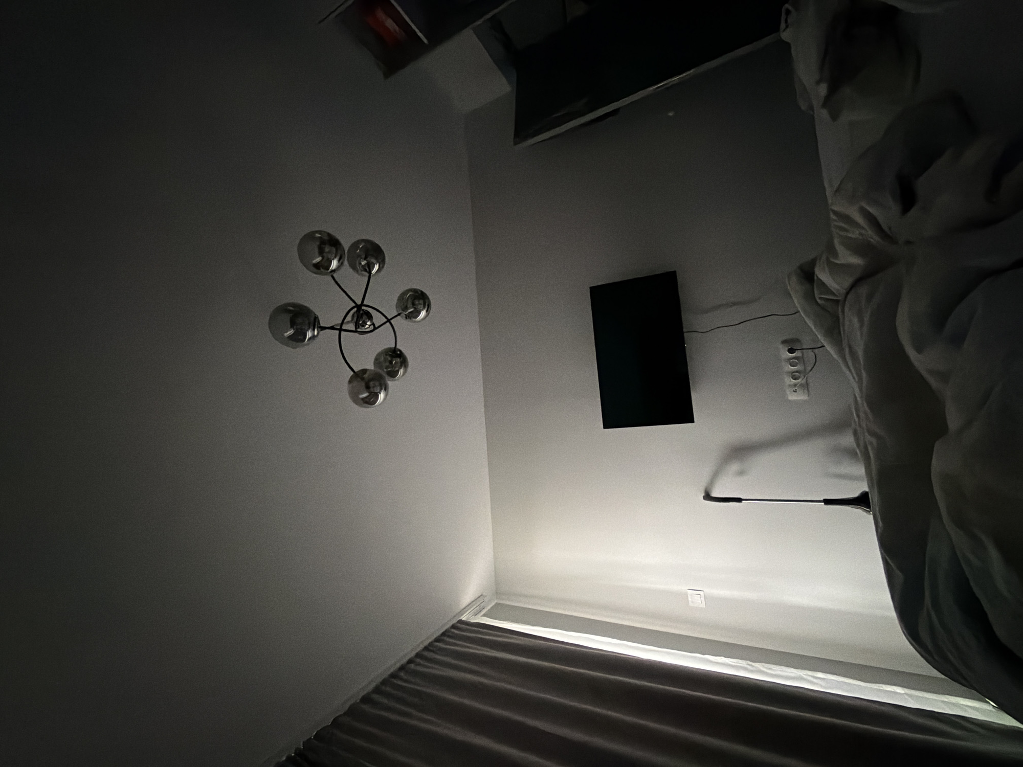 A low-angle shot of a bedroom interior. The walls are painted a light grey. A modern sputnik-style chandelier is mounted on the left wall. A flat-screen television is mounted on the wall to the right. A multi-outlet power strip is mounted below the TV. Bed curtains are visible at the bottom of the frame. A partially visible lamp is to the right, with bed sheets and comforter. The location is likely in Plovdiv, Bulgaria, but this cannot be confirmed solely from the image.