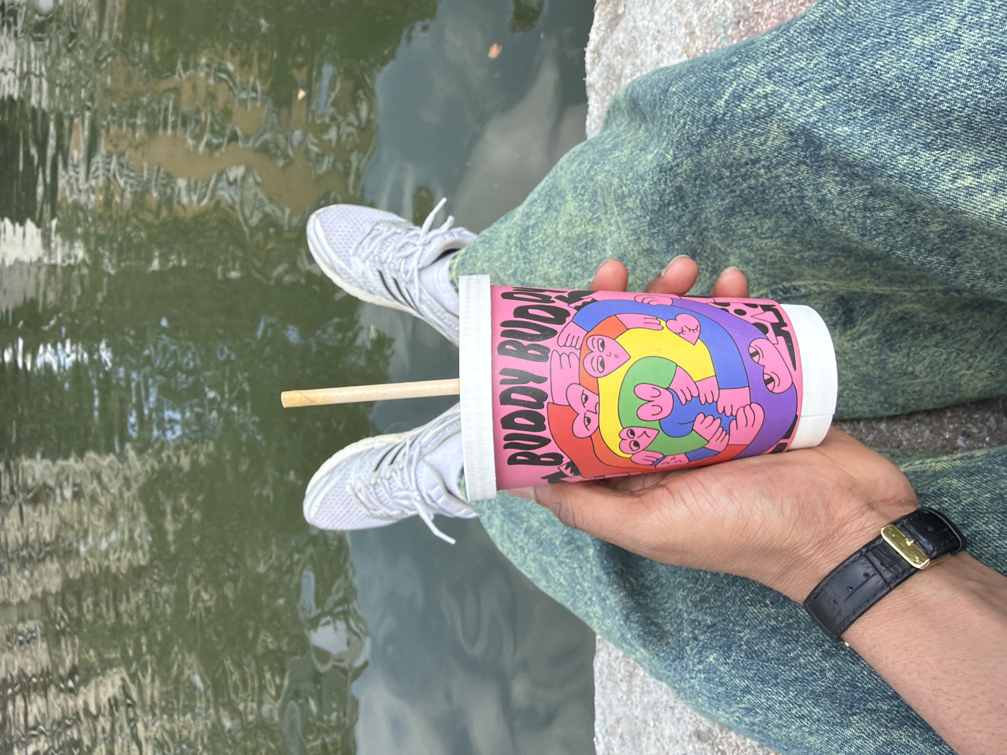 A person sits with legs outstretched in front of a body of water, possibly a canal or pond, wearing blue and green patterned jeans and white sneakers. The person is holding a brightly colored cup with the words "Buddy Buddy" printed on it, and a wooden stick is inserted into the cup. A black watch with a gold buckle is visible on the person's wrist. The water reflects buildings and trees, suggesting an urban setting, and based on the style of the clothing and the drink, the location might be in Paris, France.