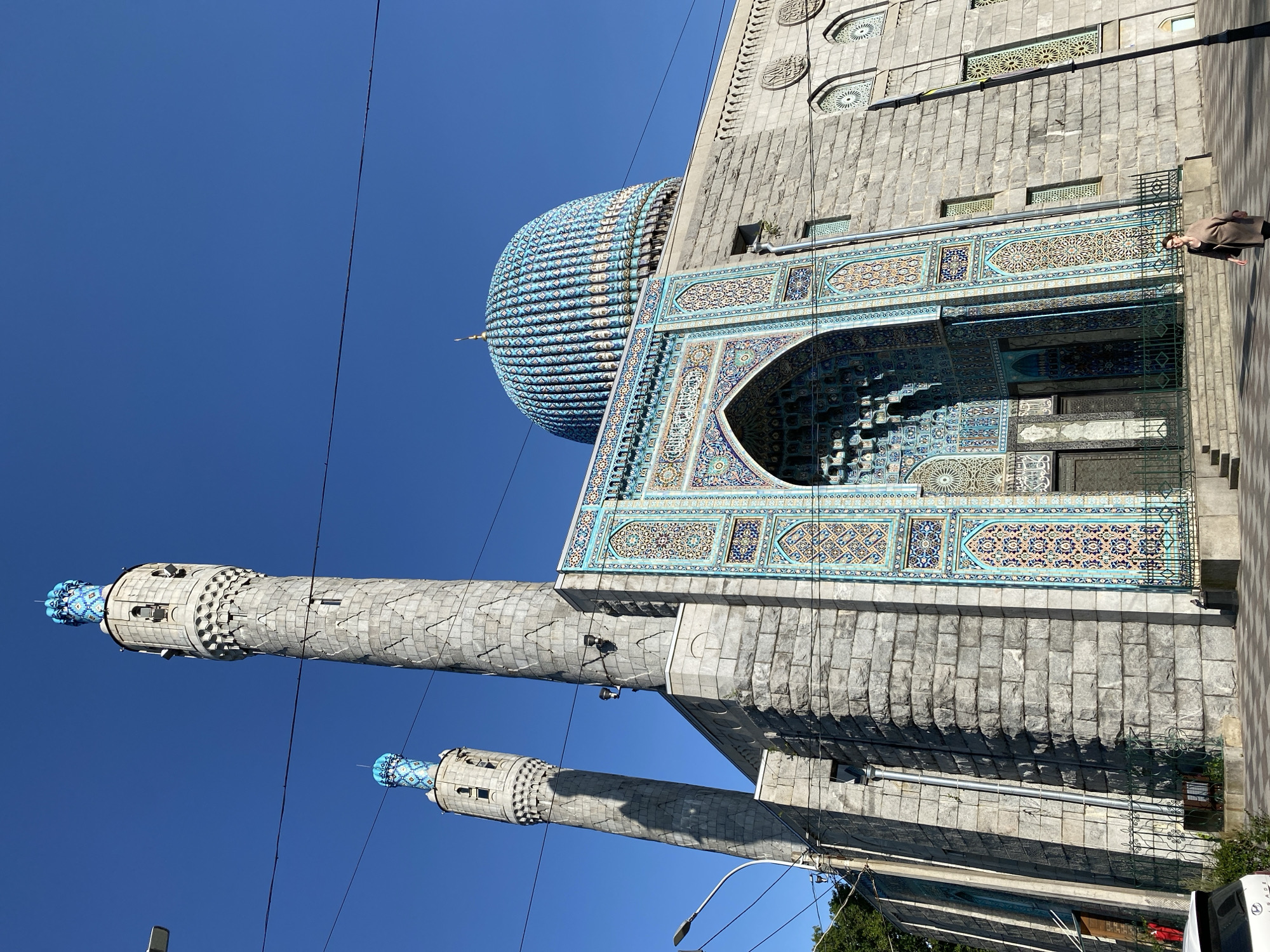 The image shows the Saint Petersburg Mosque, a large Islamic place of worship located in Saint Petersburg, Russia. The mosque features a prominent blue dome and two tall, grey minarets topped with blue decorative elements. The building is constructed with large stone blocks, and the entrance is adorned with intricate blue and turquoise mosaic tiles. In front of the mosque, a woman wearing a light brown coat walks on a paved area. The sky is a clear, bright blue, and power lines are visible across the top of the image. A street light stands to the left, and a portion of a white car is visible on the far left.