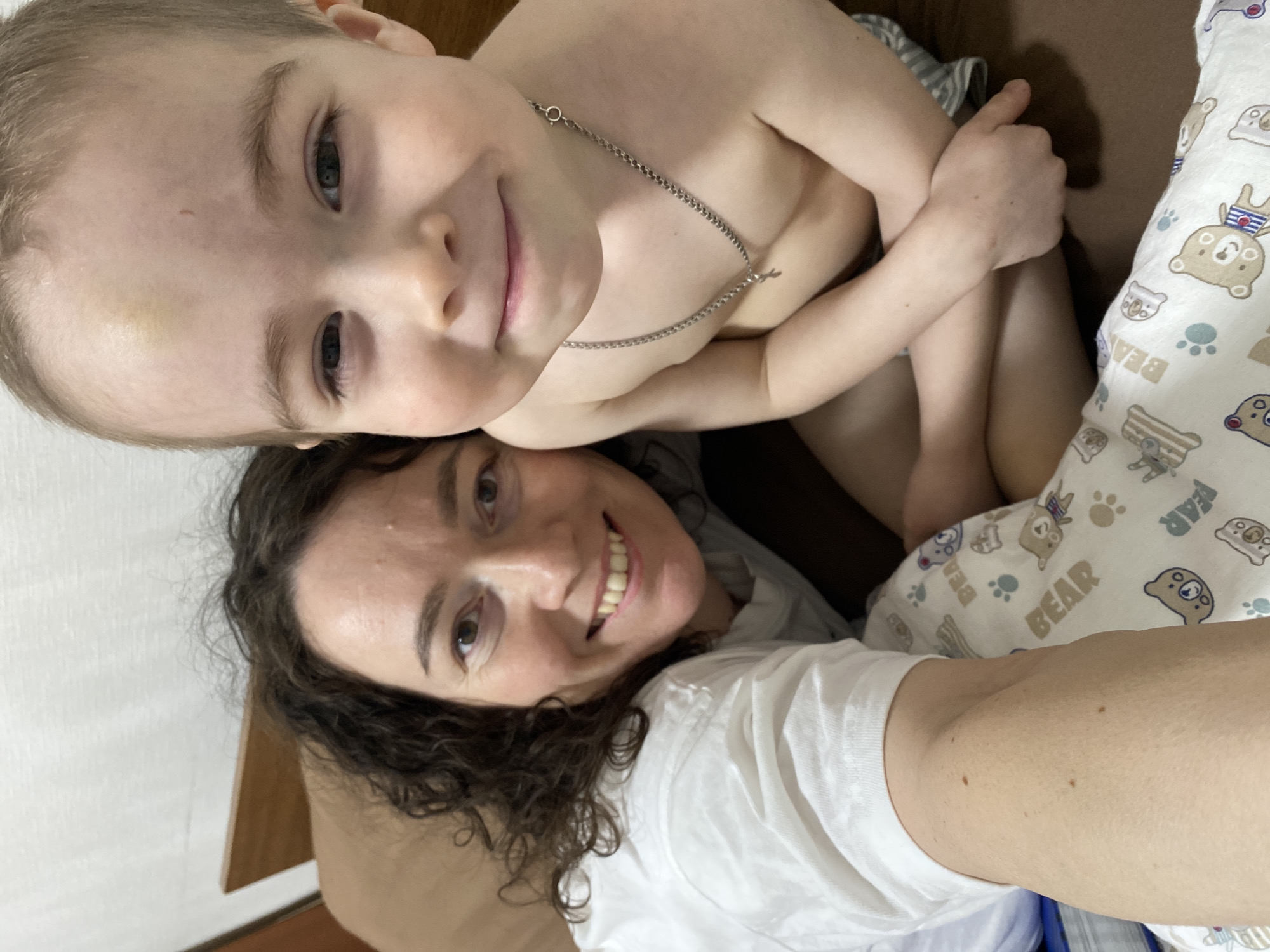 A selfie shows a woman with curly brown hair wearing a white t-shirt and a young child on top of her. The child is shirtless and wearing a silver necklace. The background includes a bed with a pillow covered in a cartoon bear print. The location is Ольховка, Russia.