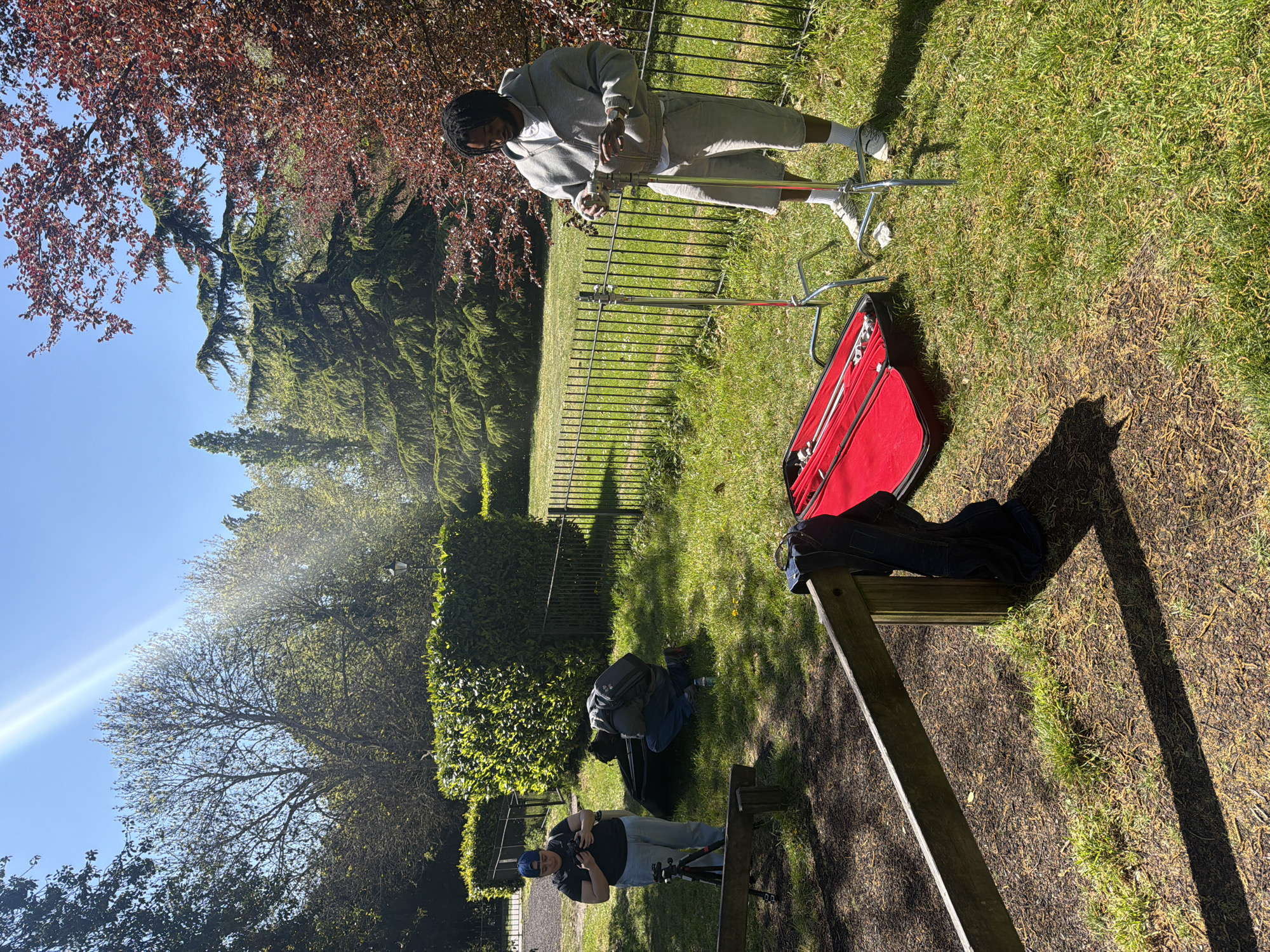 The image depicts an outdoor scene in what appears to be a garden or park. A person wearing a grey tracksuit and white socks stands on the grass near a metal film light stand. A red case, possibly containing film equipment, is open nearby. In the background, there is a metal fence, trees, and bushes. Another person wearing a blue hat, black shirt, and jeans is crouched down holding a camera, seemingly filming or photographing the first individual. Other film equipment lies on the ground, including dark clothing and a bag. A wooden structure, possibly part of park seating, is in the foreground, casting a shadow. Based on the architecture and foliage observed in the image, the location is potentially within the city of London, United Kingdom.