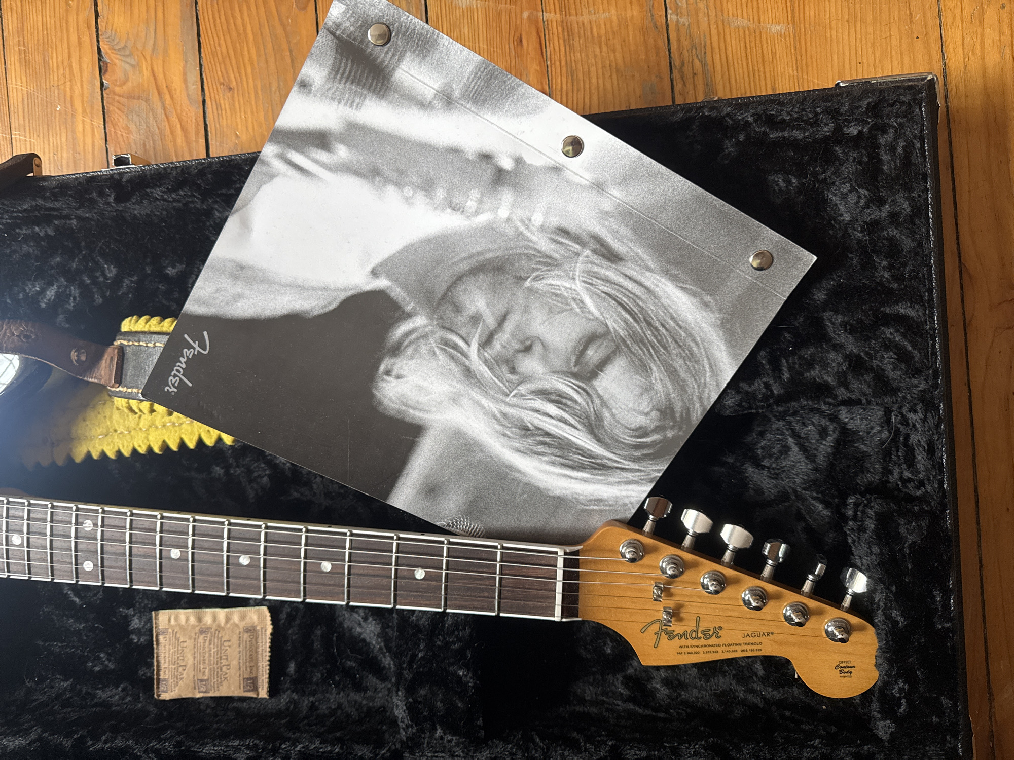 An overhead shot displays a Fender Jaguar electric guitar resting in its open black velvet-lined hard case on a wooden floor. A black and white print, possibly a poster or cover art, lies diagonally across the guitar's body and case. This print features the blurry, close-up face of a person with long hair, appearing to be lying down, and is fastened at the top with two visible metal studs.
The guitar's neck and headstock are prominent, showcasing the "Fender" logo and "JAGUAR" model name. Below the model name, text reads "WITH SYNCHRONIZED FLOATING TREMOLO" and lists several patent numbers: "PAT 2.960.000 2.972.923 3,143.026 065 186.820". The headstock also features "OFFSET Contour Body". Guitar tuners are visible along the headstock. A section of a guitar strap, yellow and black, is visible near the base of the neck, with "Fender" text partially discernible.
Below the guitar's fretboard, resting on the case's velvet lining, is a small, rectangular brown desiccant packet. "UNIT PAK" is printed multiple times on this packet. The wooden floor serves as the background for the guitar case and its contents. No specific activity is depicted beyond the arrangement of these items, suggesting a display or photograph of personal belongings.