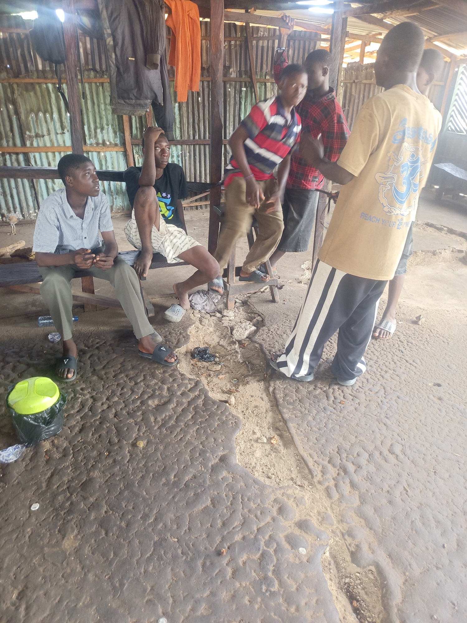 A photograph taken in Akwanga, Nigeria, shows a group of young men gathered in an open-air structure. Two men are seated on a wooden bench on the left side of the frame, with one wearing a vertically-striped shirt and the other in a black T-shirt and patterned shorts. To the right, three other men are standing. One is wearing a red and white striped shirt and tan pants, another in a red plaid shirt and dark shorts, and a third man is wearing a mustard-colored T-shirt and black and white striped pants. The flooring appears to be uneven and worn, with some debris scattered around. A green and black trash can sits to the left of the seated men. Clothes are hanging in the background. The environment appears informal and relaxed.