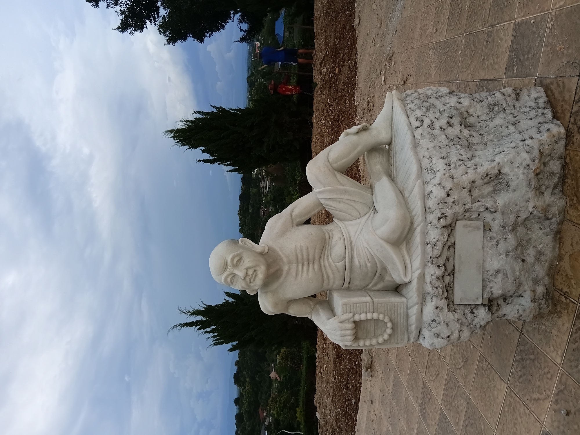A white marble statue of an elderly man is seated on a rough, unpolished stone pedestal. The statue depicts the man with a thin, skeletal frame, wearing simple robes and holding a string of large beads in his left hand. He is positioned on a tiled area, with a hilly, wooded landscape in the background. The sky is overcast and gray, suggesting a cloudy day. In the mid-ground, two figures appear to be working on the landscape; one is wearing a red top and a wide-brimmed hat, and the other is wearing a blue shirt and shorts. The location is identified as Ban Lao Phatthana, Thailand.