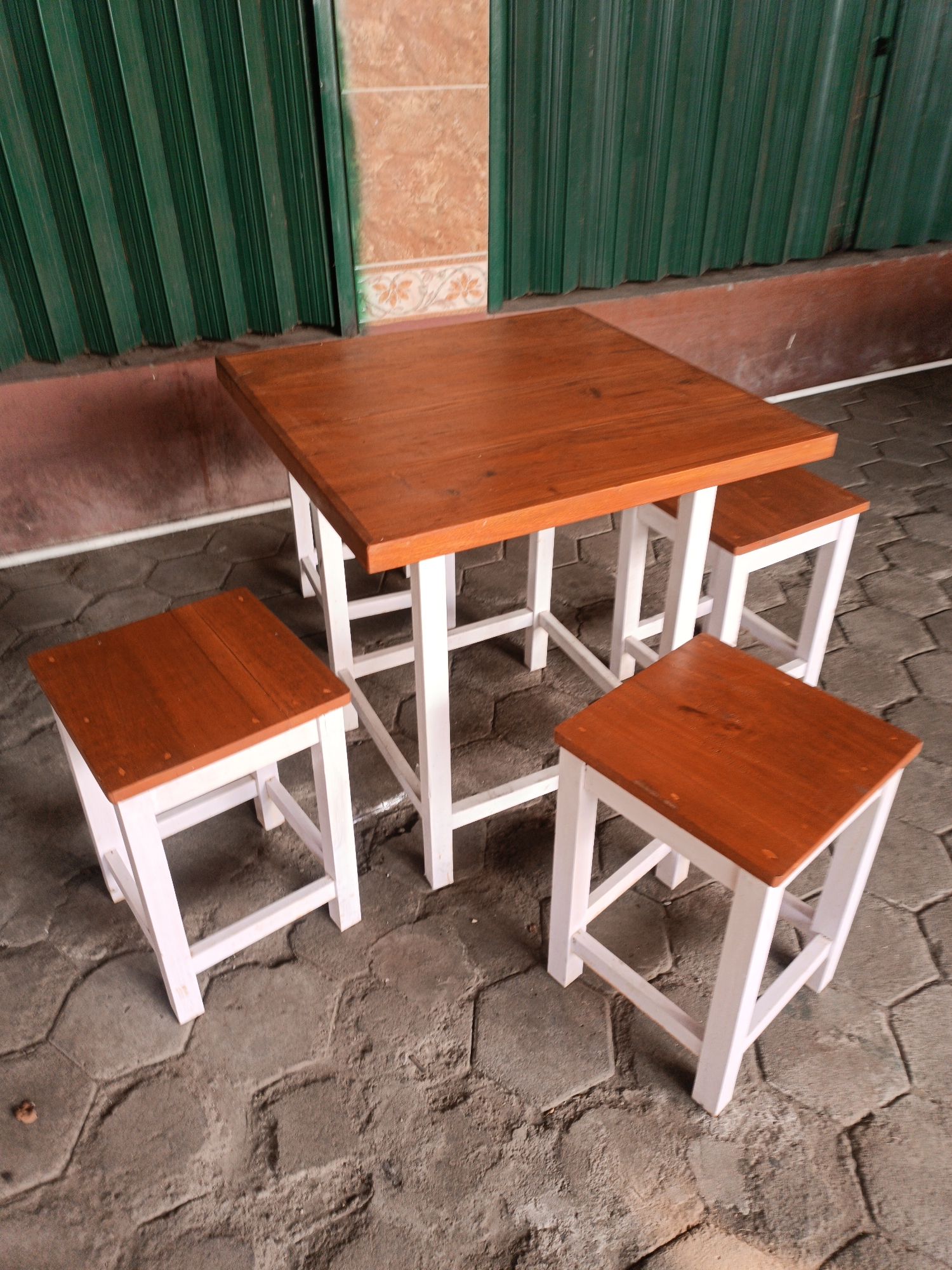 In Imogiri, Indonesia, a wooden table and four stools are set up outdoors on a patterned stone ground. The table is square and made of light brown wood with white legs. Three of the stools are visible and arranged around the table, also made of light brown wood with white legs. Behind the table and stools is a green corrugated metal door, partially open, revealing a tiled wall in shades of orange and white. The scene suggests a seating area outside a shop or establishment.