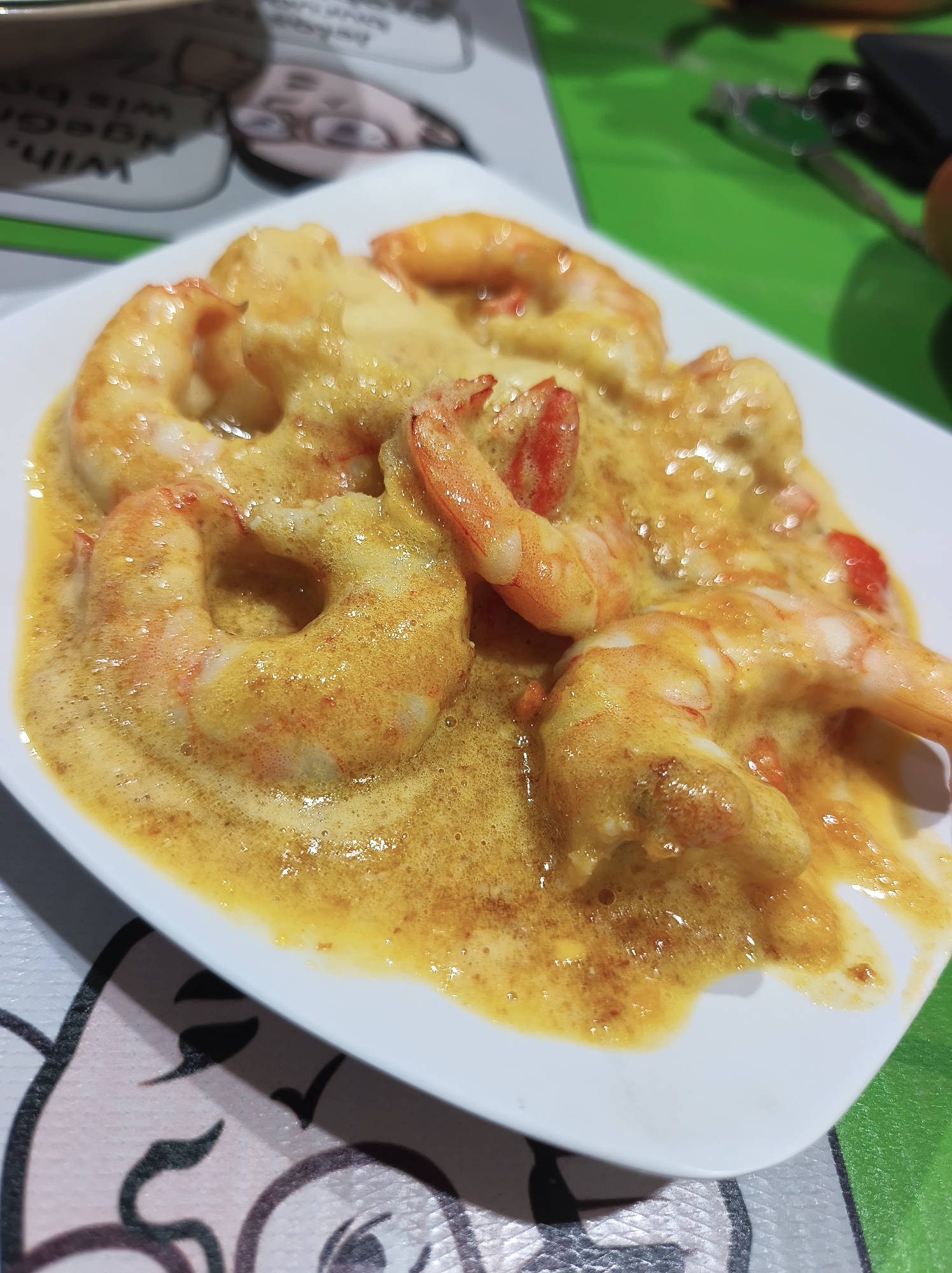 A close-up, eye-level shot shows a white rectangular plate of shrimp in a creamy yellow sauce. The shrimp are pinkish-orange and curled, with visible segments. They are coated in a frothy, textured sauce that appears to be seasoned with spices, as indicated by small dark specks. The plate is positioned on a surface with a patterned placemat and a vibrant green surface in the background. The lighting creates reflections on the sauce, highlighting its glossy texture. The background is slightly blurred, but a hint of a menu or paper with text and illustrations can be seen in the upper left. The overall scene suggests a meal being served in a casual dining setting, possibly a restaurant. The image does not contain enough information to determine its specific location in Kebonsari, Indonesia.