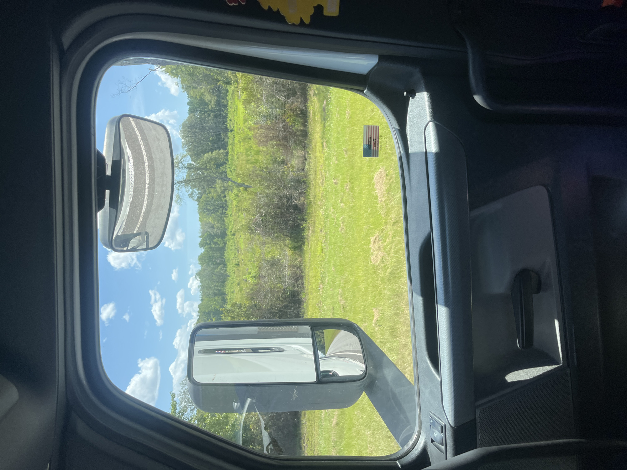The image is taken from inside a vehicle, likely a truck, looking out the window. The view is of a rural landscape with green grass and trees under a bright blue sky with scattered white clouds. A large side mirror of the truck is prominently featured on the left side of the frame, reflecting the road ahead. A smaller secondary mirror on the side mirror shows a reflection of the road curving to the right. A small American flag sticker is visible on the exterior of the truck door. The interior of the truck is visible in the foreground, including a door handle and part of a seat. The scene appears to be in Riceboro, United States, judging by the context.