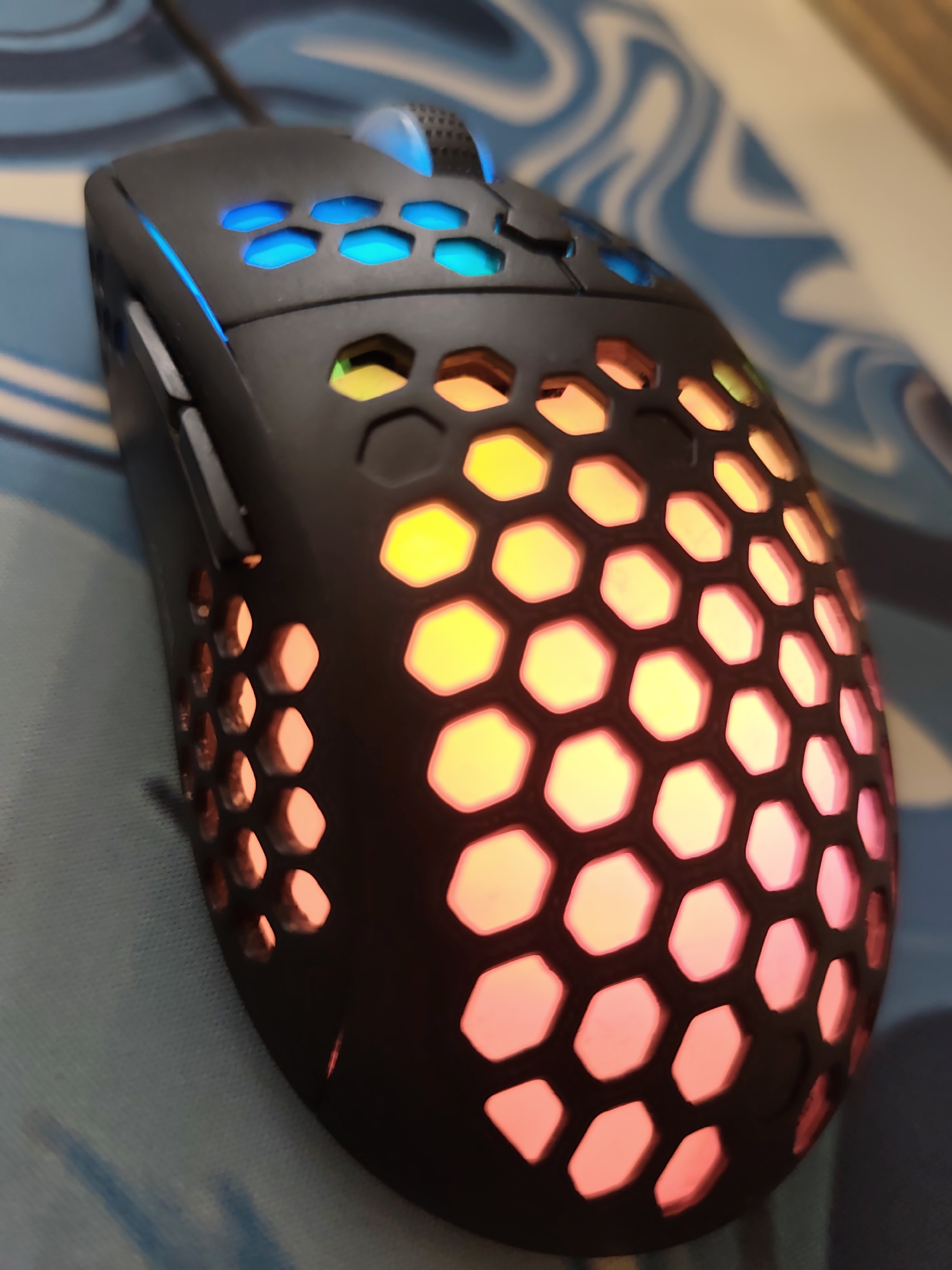 A black gaming mouse featuring a perforated honeycomb shell is depicted, illuminated internally with an RGB gradient. The front portion near the scroll wheel and under the main click buttons emits blue light. The middle section of the honeycomb pattern glows with yellow and orange light, transitioning to pink and red light towards the rear. The mouse includes a translucent blue scroll wheel and two dark grey side buttons on its left flank. A braided cable extends from the front of the device. The mouse rests on a light blue surface adorned with an abstract, wavy white and darker blue pattern, indicative of a mouse pad. The background is out of focus, showing a continuation of the patterned surface and a light brown or beige vertical surface. No individuals are present in the image. The image contains no discernible information to ascertain its geographic location, thus the assertion of "Pazardzhik, Bulgaria" cannot be verified based on visual evidence.