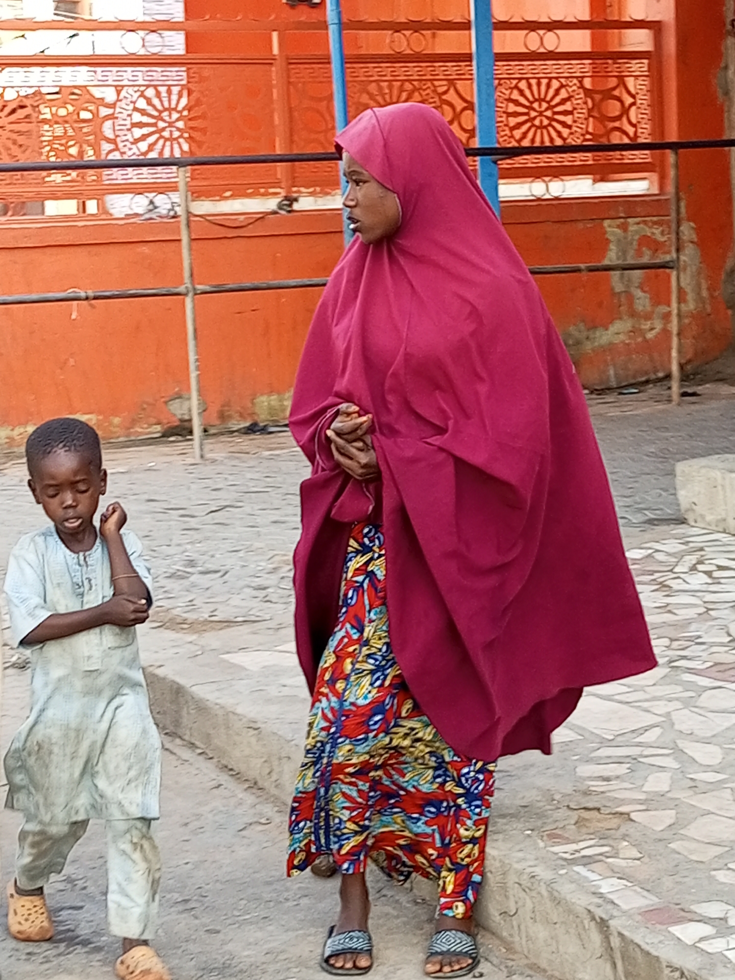 In the city of Arawa, Nigeria, a young boy and a woman are depicted walking on a paved street. The boy, appearing to be around seven years old, is wearing a light-colored, long-sleeved tunic and trousers, with tan slip-on shoes. He is looking downwards with his arms crossed. The woman is dressed in a vibrant, patterned skirt with shades of red, blue, and yellow, and she is wearing a dark pink hijab that covers her head and body. Her hands are clasped in front of her. They are walking on a street with an orange wall and intricate metalwork in the background. The ground is a mosaic of light and dark stones.