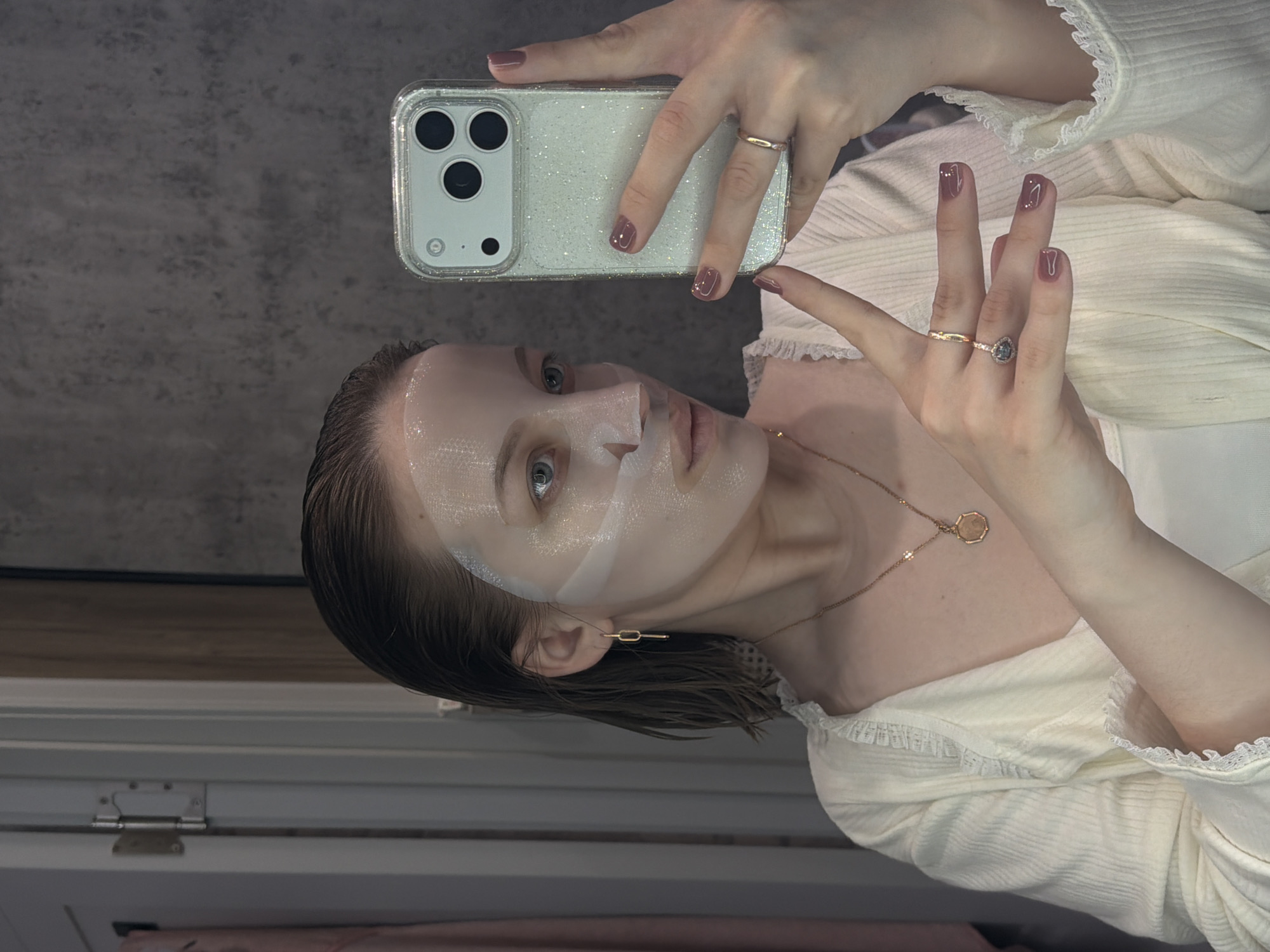 A woman with light skin and wet, short, light brown hair is taking a selfie. She has a white sheet mask on her face. She is wearing a light-colored blouse with lace trim and a gold necklace with an octagonal pendant. Her nails are painted a shiny dark pink. She is wearing two gold rings on her right hand. She is holding a white smartphone with a sparkly case. The background is a gray wall and a white cabinet. The location is not discernible from this image.