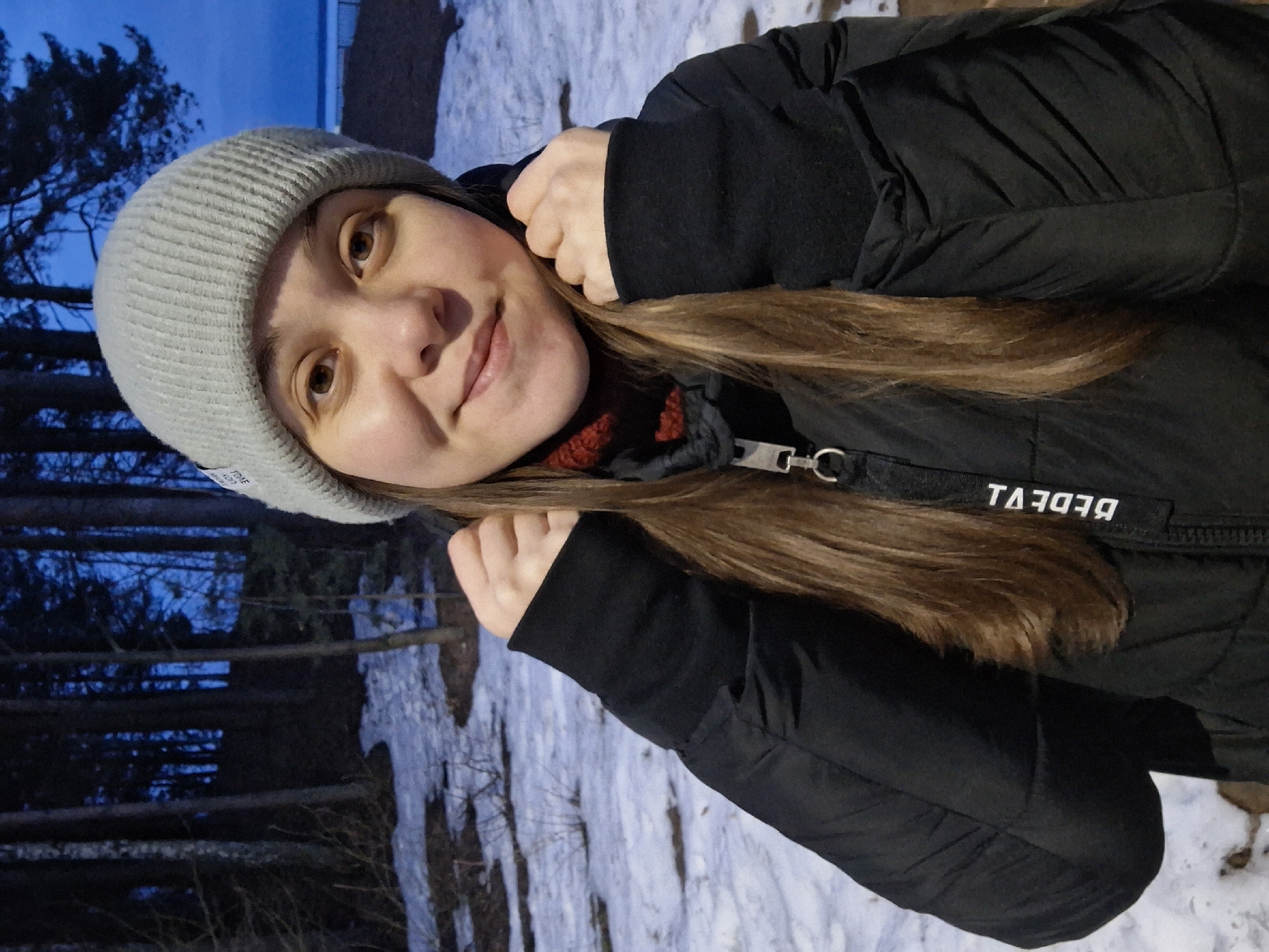 A portrait of a woman with light brown hair and brown eyes, wearing a gray beanie and a black winter jacket with a visible label on the front. She is smiling slightly. The background shows a snowy forest with dark trees, suggesting it's dusk or early evening. The photo was taken in the city of Chaikovsky, Russia.
