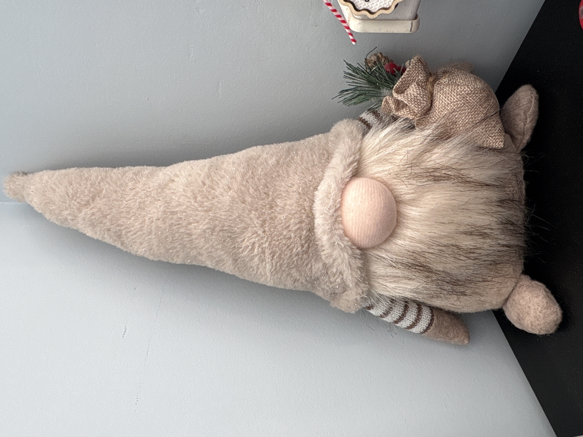 An indoor, close-up photograph captures a plush gnome figure positioned horizontally against a light grey wall, resting on a dark surface, likely a shelf or cabinet. The gnome, predominantly light beige and tan, occupies the central and left portions of the image.
Its tall, conical hat, made of soft, fuzzy fabric with subtle glittery threads, extends towards the left. A prominent, round, peach-colored nose protrudes from beneath the hat. Below the nose, a voluminous beard of light brown and grey faux fur cascades towards the right. Adorning the upper right side of the gnome's head is a small burlap bow, accented with a miniature green pine sprig and tiny red berries. Further to the right, a small, rounded, beige ear-like protrusion is visible. A striped arm or leg, featuring brown and beige bands, extends downwards from the gnome's body, while another rounded, foot-like appendage rests on the dark surface to the far right.
The background consists of a textured, light grey wall. In the top right corner, partially visible above the gnome's head, are a white and red striped candy cane and a fragment of a wooden decorative item, possibly part of a miniature house or sign. No specific activity is depicted, as the image is a still life of a decorative object. There is no visible text, and the time of day, weather, or emotions are not discernible.
