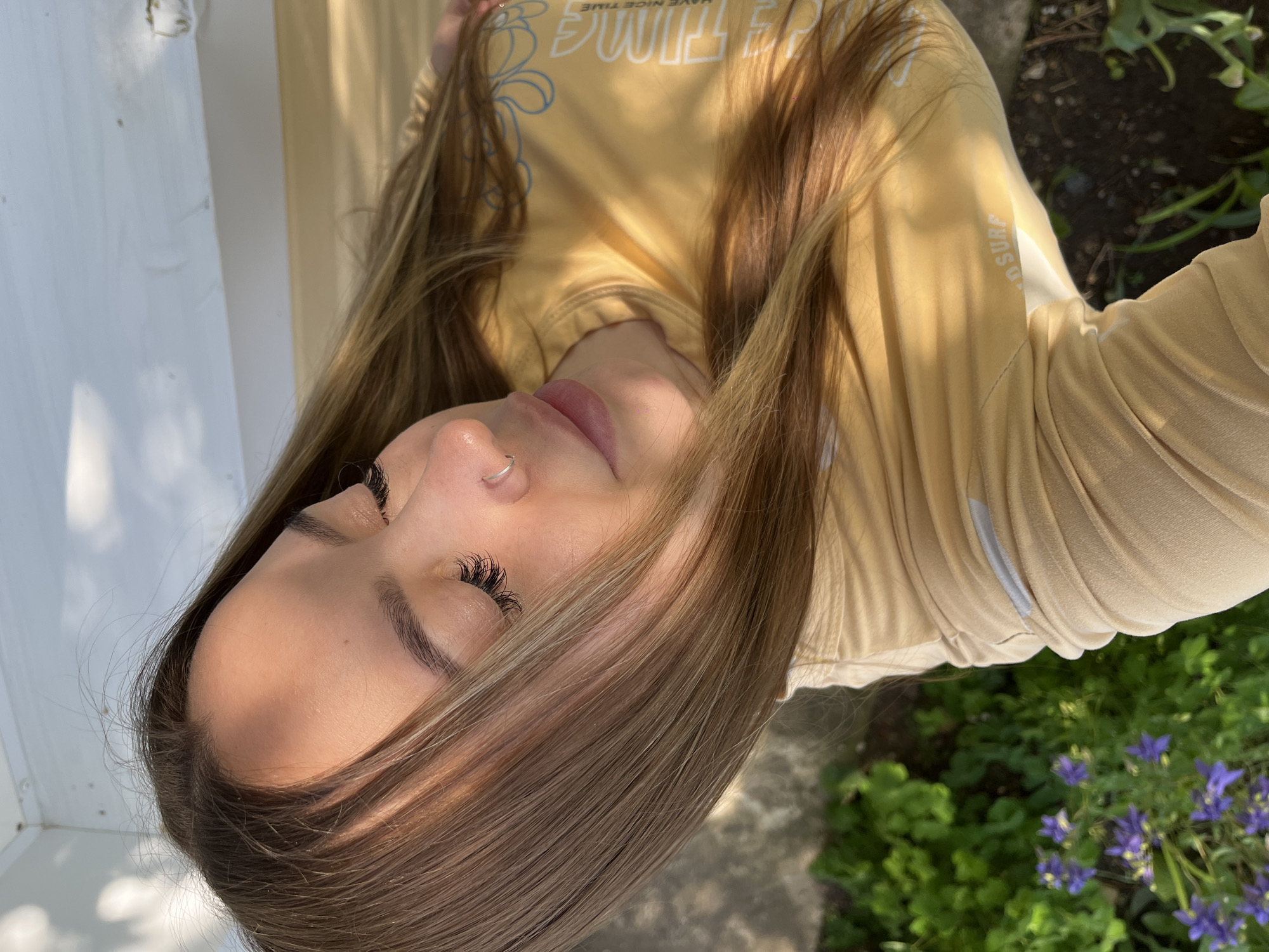 A young woman is lying down outside. Her hair is long and brown. She is wearing a yellow shirt. There are plants and flowers in the background. The location is not determined to be the city of Novopokrovskaya, Russia.