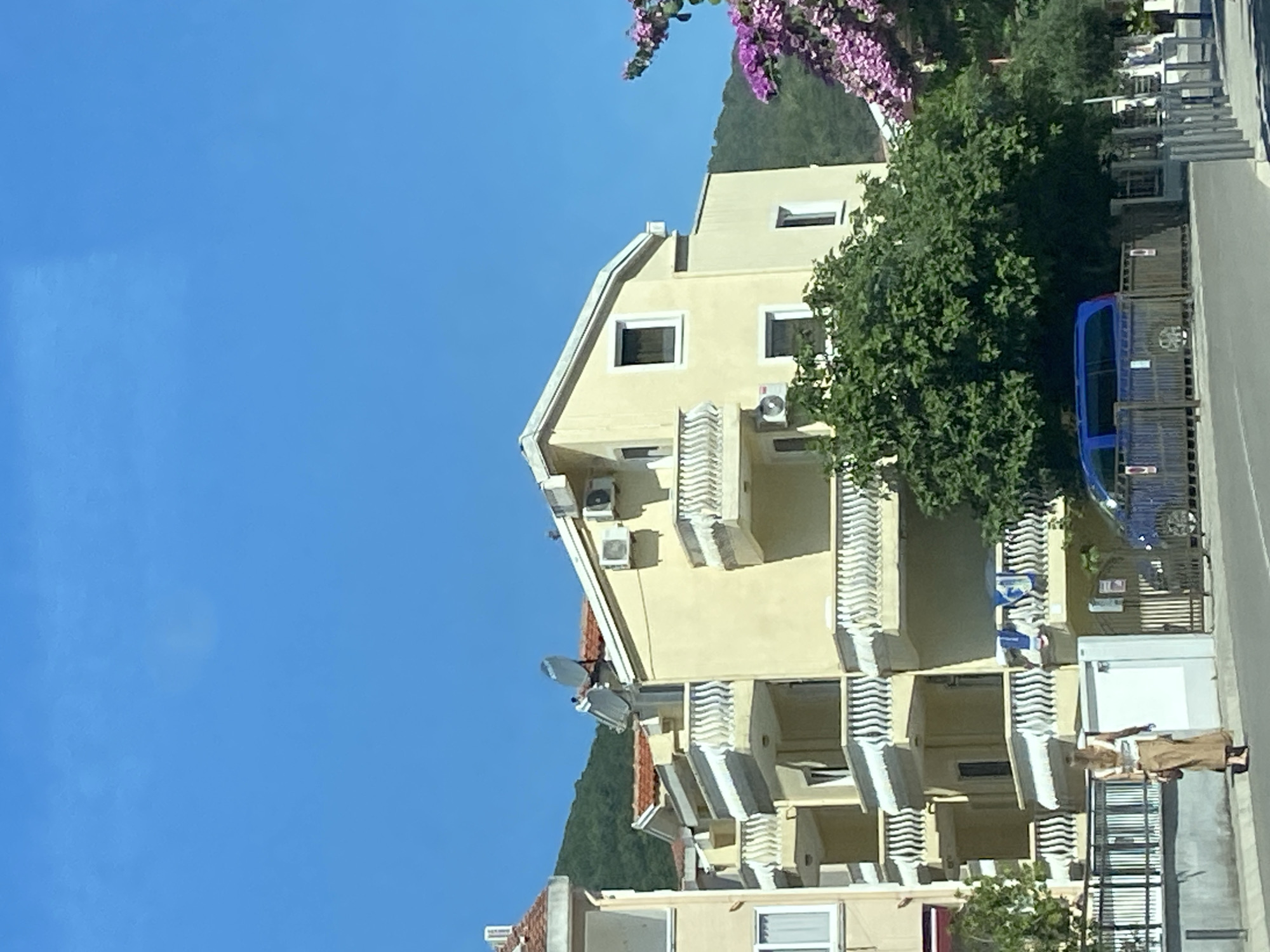 A light yellow building with multiple balconies and satellite dishes on the roof stands under a clear blue sky. A lush green tree is in front of the building. To the right, a tree with bright purple flowers is visible. A woman wearing a long, tan skirt and a white top walks away from the camera on a sidewalk. A blue van is parked behind a wooden fence. The scene appears to be in a residential area of Budva, Montenegro.