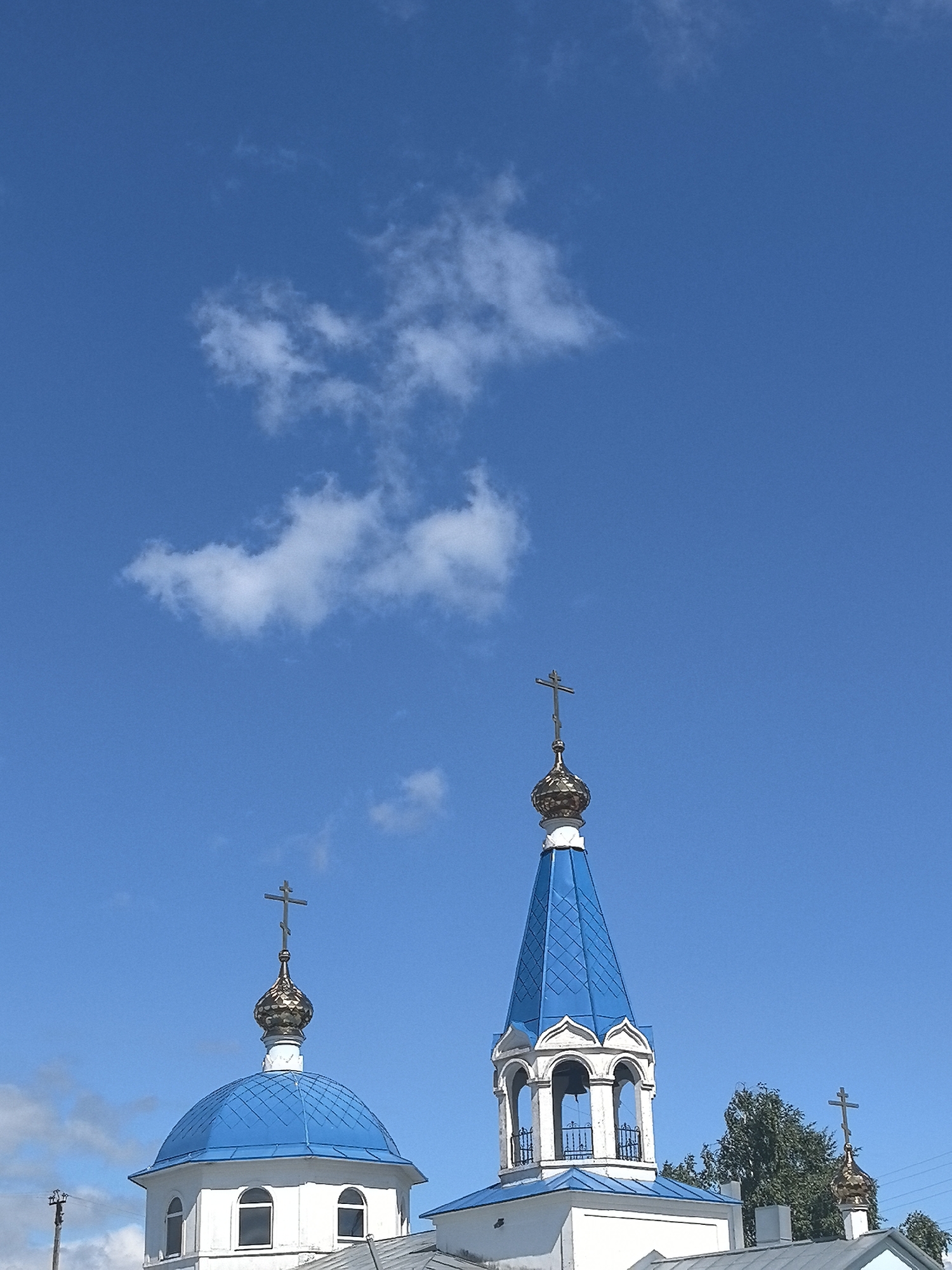 The image shows the rooftops of several structures in Noviy, Russia, under a bright blue sky with sparse white clouds. Dominating the scene are the blue, onion-shaped domes of what appear to be Orthodox churches. Two prominent domes are visible, one with a cross on top, and another that is taller and more conical, also topped with a cross. In the background, trees and some electrical poles are visible, suggesting an outdoor, possibly semi-urban or rural, environment. The architecture indicates a religious setting typical of Russian Orthodox churches.
