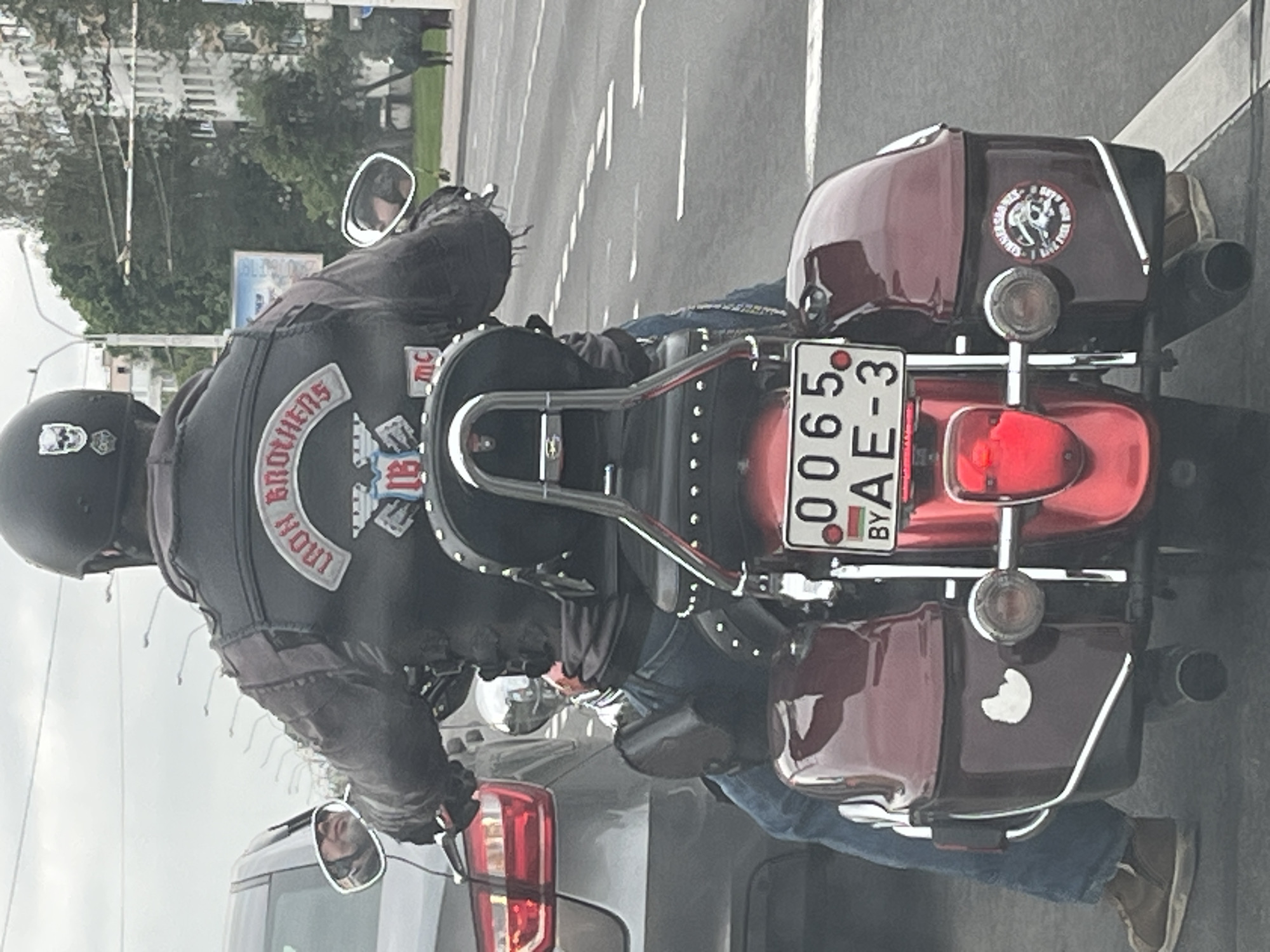 The image displays a motorcyclist from a rear perspective, stopped on an urban or suburban road, likely in Minsk, Belarus, as evidenced by the "BY" country code on the license plate. The scene is set during the daytime, possibly under an overcast sky.
The central subject is a person wearing a black helmet adorned with a white skull decal, a black leather vest with various patches, and blue jeans. Notable patches on the vest include a top rocker reading "BROTHERS" and a central emblem that appears to say "IRON MC" with a skull design, accompanied by a smaller "MC" patch below it.
The motorcycle is a dark red or maroon cruiser model, featuring chrome accents, hard saddlebags, and a chrome backrest. The Belarusian license plate, displaying "0065 AE-3 BY," is clearly visible. The right saddlebag sports a circular sticker with a skull and partially legible text that seems to include phrases like "I RIDE SO I LIVE THE WILD ONE." Another lighter-colored decal, possibly a crescent shape, is present on the left saddlebag.
The motorcycle is positioned behind a car, whose rear light and part of its body are visible in the foreground, indicating traffic. The background includes trees, buildings, and a paved road with white lane markings. The rider's face is not visible.