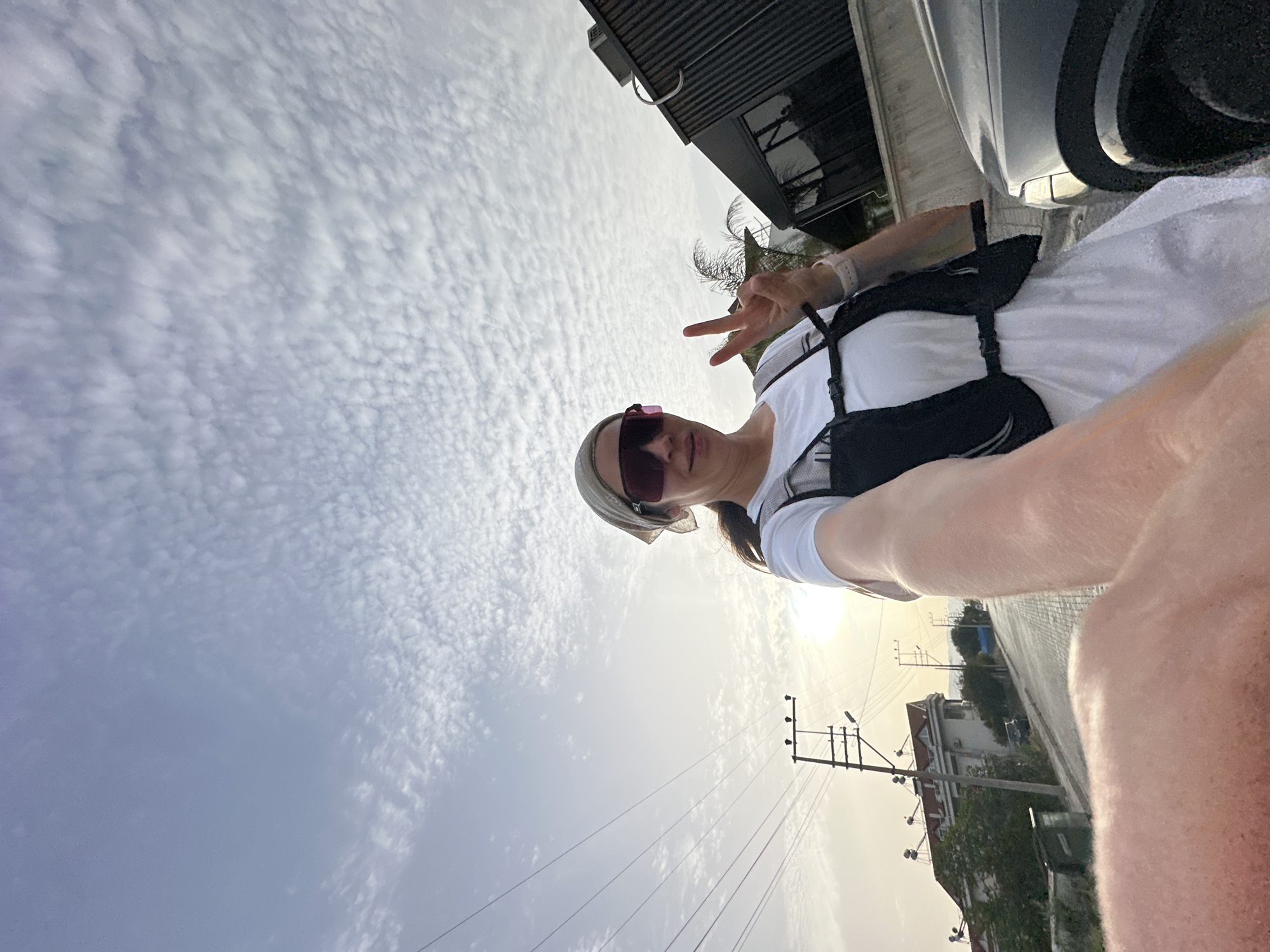 The photograph depicts a woman taking a selfie. She is wearing sunglasses and a headscarf, a white top, and a black harness or backpack. She is holding up two fingers in a peace sign. Behind her is a partly cloudy sky, and below her is a street scene including buildings, power lines, and trees. The location is Fethiye, Turkey.