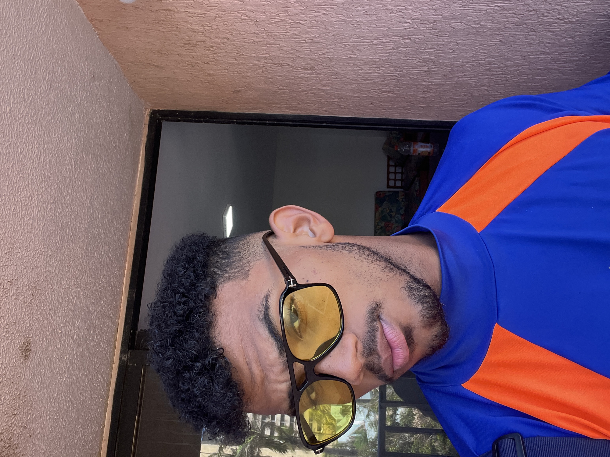 A man with dark curly hair, a mustache, and a goatee is pictured outdoors, likely on a balcony in Porlamar, Venezuela. He is wearing yellow-tinted sunglasses and a blue long-sleeved shirt with orange accents. The background shows a textured wall, a door with glass, and glimpses of greenery and possibly buildings outside.