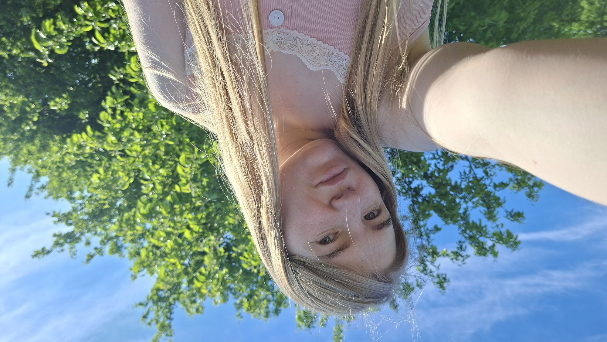 This media file is an inverted portrait-style selfie taken outdoors, featuring a person with long blonde hair, fair skin, and light-colored eyes, looking directly at the camera with a neutral expression. The individual is wearing a light pink ribbed top with a white lace trim around the neckline and a single white button visible on the garment. Their arm is visible on the right side of the frame.
The background consists of vibrant green tree foliage and a clear, bright blue sky with some wispy white clouds. The perspective is from a low angle, looking upwards, as if the subject is lying down and gazing towards the sky, or the camera is held below them. This orientation places the subject's head at the bottom-center of the frame, with their hair flowing upwards, and the sky and trees forming the upper half of the image.
The lighting indicates bright daylight, suggesting a sunny day. There are no specific activities depicted other than posing for the photograph. No visible text is present in the image. The scene conveys a serene and natural outdoor moment. The location context provided is Большой Букор, Russia.