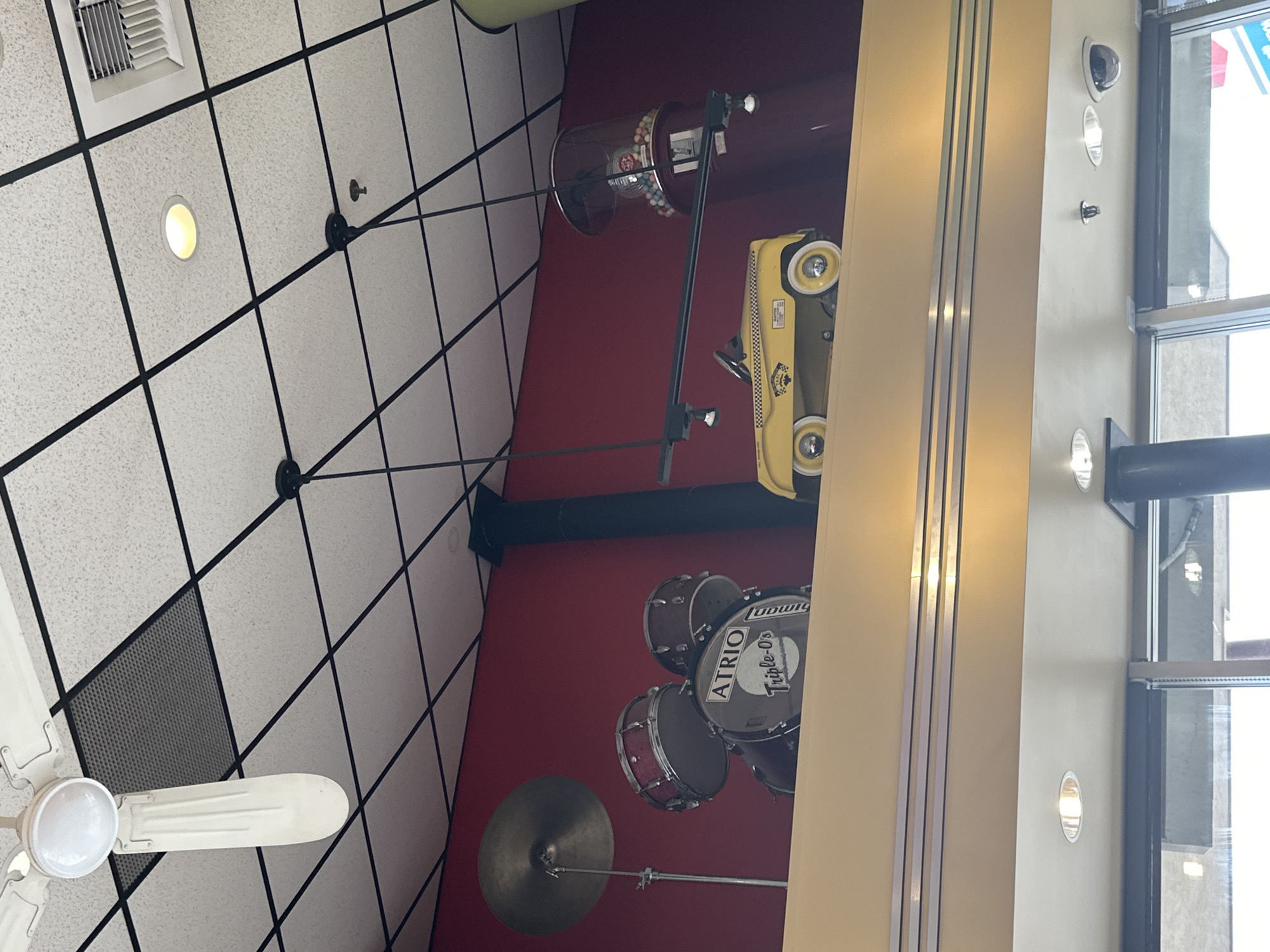A ground-level, interior view shows ceiling tiles with inset lighting and ceiling-mounted objects attached to a red wall. The objects include a drum set with "ATRIO" branding on the drum head, a cymbal, a yellow toy taxi cab, and a candy machine. To the right, a window column shows multiple inset lights. In the bottom left corner is a white ceiling fan. Based on the business logo visible in the window, this image was taken at a building within Township of Langley, Canada.