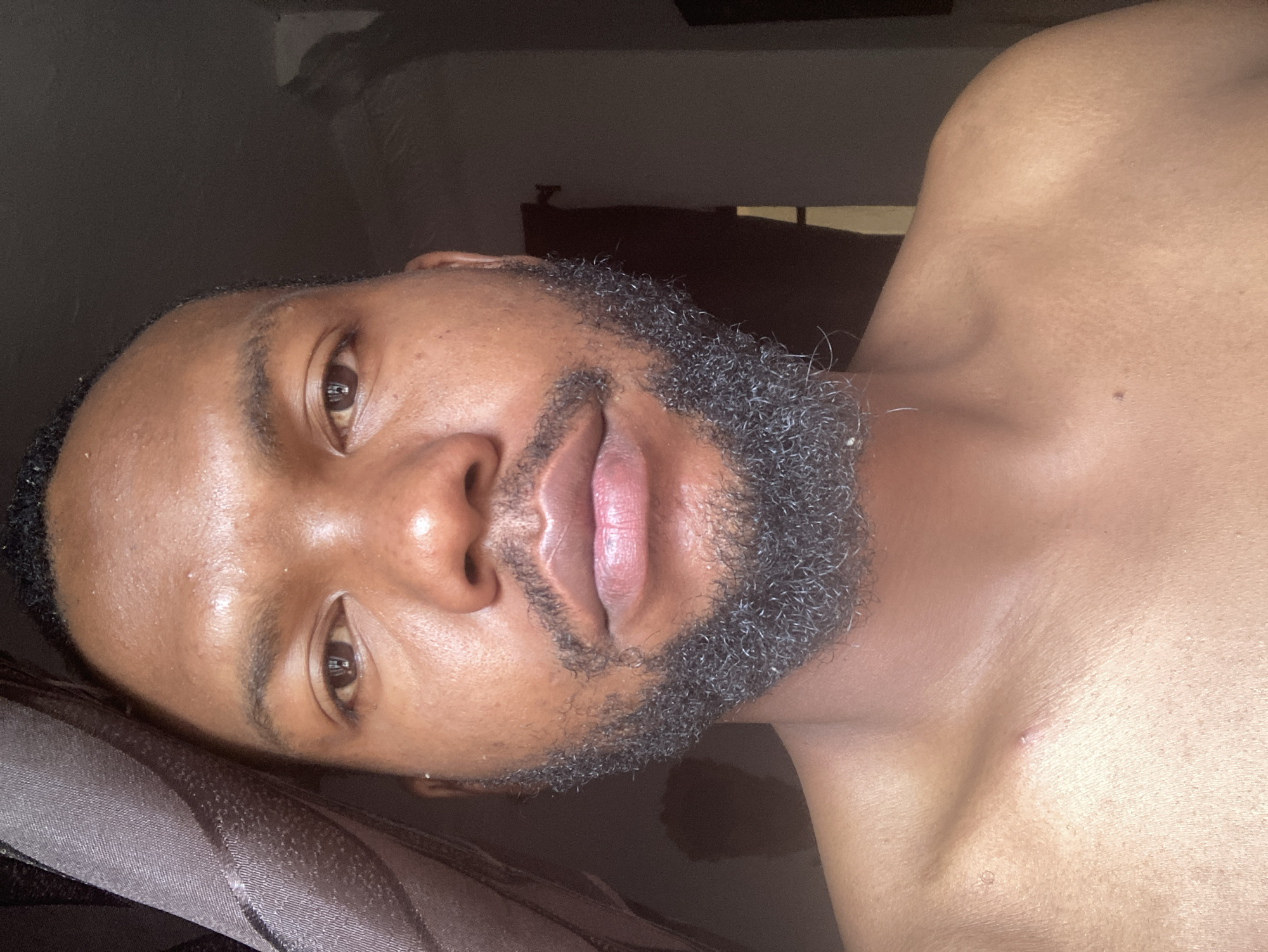 A close-up, selfie-style portrait of a man with dark skin and a short, dark, curly beard. He is lying down, possibly in a bed, as indicated by the visible pillow or bedding in the lower left corner. His torso is bare. The background is dark and indistinct, with a window visible in the upper center of the frame. No information in the image specifically indicates that this photo was taken in Jos, Nigeria.