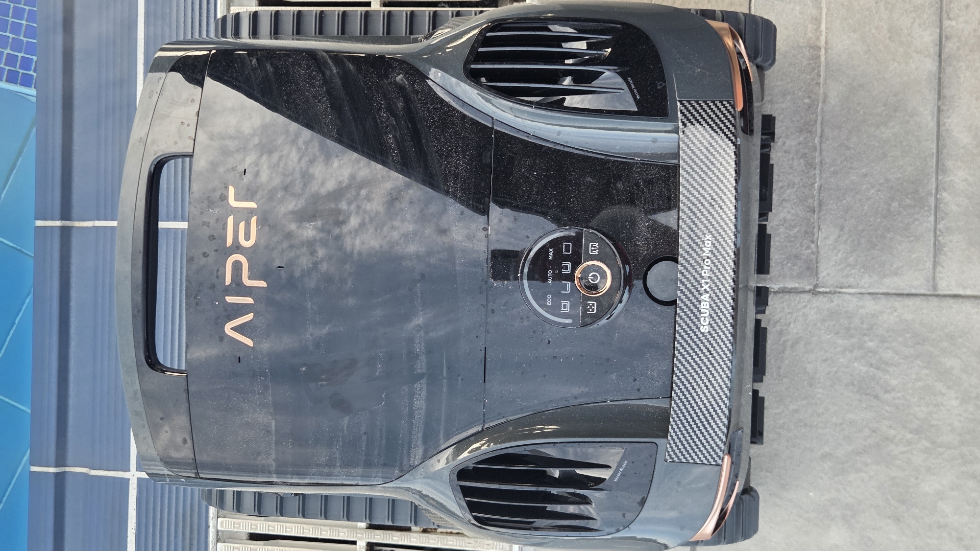A top-down, full shot shows a black, robotic pool cleaner with copper accents and the "AIPER" logo on its upper surface. The cleaner is positioned on a tiled surface, possibly a patio, next to a blue tiled area, likely a pool. The cleaner has a handle at the top, ventilation grilles on its sides, and a control panel in the center with buttons labeled "ECO," "AUTO," and "MAX," along with a power button. The words "SCUBA XI Pro Max" are visible on a decorative band near the bottom of the cleaner. The environment appears to be outdoors.