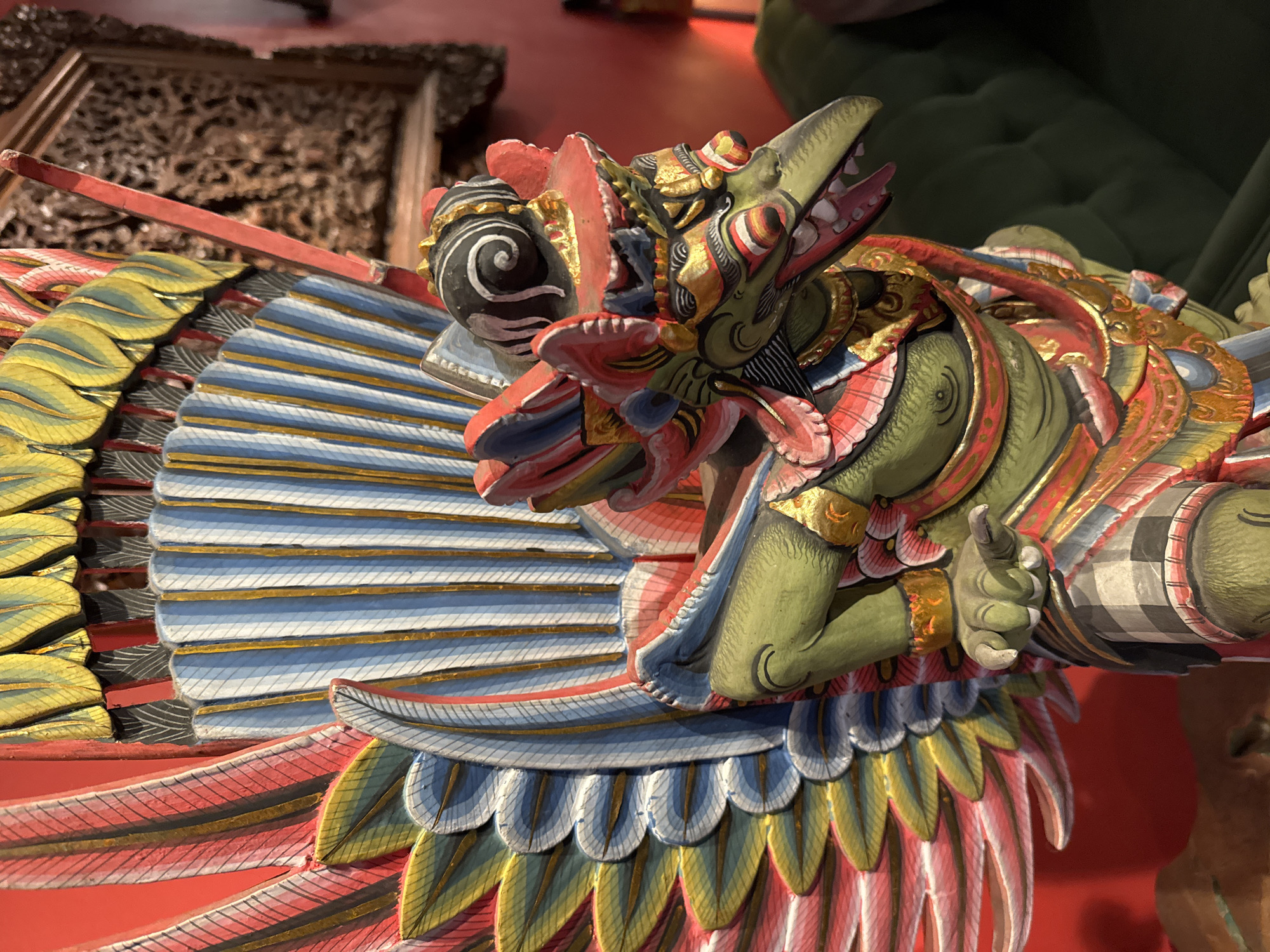 A carved wooden effigy, likely a Garuda or similar mythological bird-like creature, stands prominently. The effigy is vibrantly painted with intricate details, featuring a green body, a long beak with visible teeth, elaborate feathered wings in red, yellow, blue, and white, and decorative gold accents. The creature wears a patterned lower garment resembling a sarong or skirt. The background consists of a red wall on the left and a blurry, dark green upholstered surface on the right, possibly a sofa. The setting appears to be indoors, suggestive of a shop or decorative display, and aligns with the typical artistic style found in Kuta, Indonesia, known for its traditional wood carvings and religious iconography.