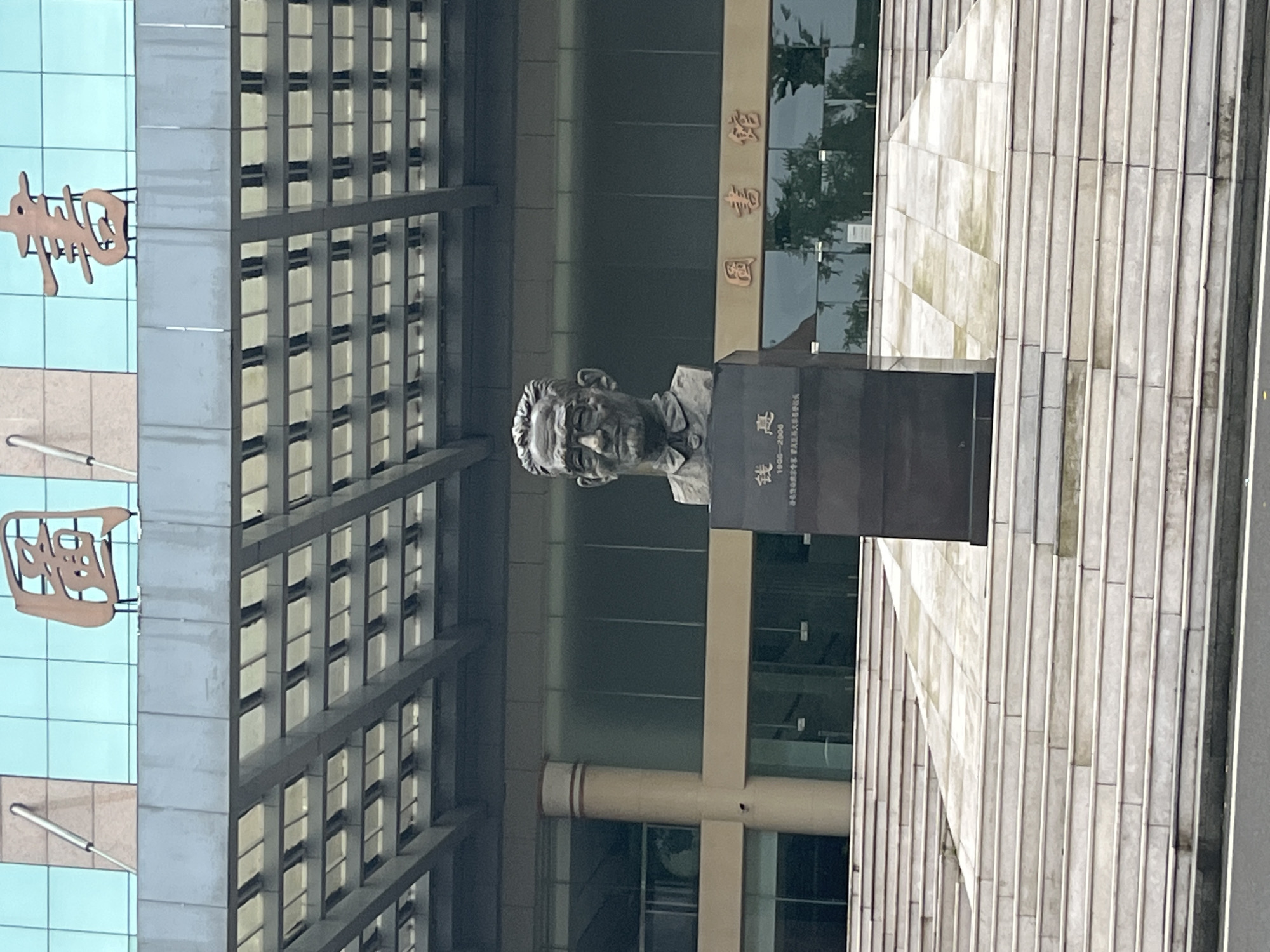 This image presents the front exterior of a library building, situated in Yuzhong District, China. A dark bronze bust of a man is centrally positioned in the foreground, mounted on a tall, dark rectangular pedestal. The bust depicts a male figure with short, styled hair, wearing glasses and a collared shirt with a tie. The pedestal bears Chinese characters "钱穆" (Qian Mu) and the years "1895-2002" below the name, followed by smaller Chinese text translating to "Famous Chinese historian and educator."
The library building behind the bust is a modern structure with large glass panels and light-colored horizontal bands. Prominently displayed on the upper facade are three large, relief-style Chinese characters in a reddish-brown hue, spelling "图 书 馆" (Tú Shū Guǎn), which translates to "Library." Below these characters, a complex, multi-tiered structural overhang with a grid pattern provides shade. The lower section of the building features extensive glass windows that reflect the surrounding environment, including what appears to be trees. A cylindrical column in a light beige color is visible on the left side of the entrance.
A wide, multi-level staircase made of light-colored stone steps leads up to the library entrance from the immediate foreground. An inclined ramp section is integrated into the right side of the staircase, adjacent to the bust's pedestal. The lowest part of the image shows a strip of dark asphalt, indicating a road or paved area at the base of the steps. The sky appears overcast, suggesting a cloudy day.