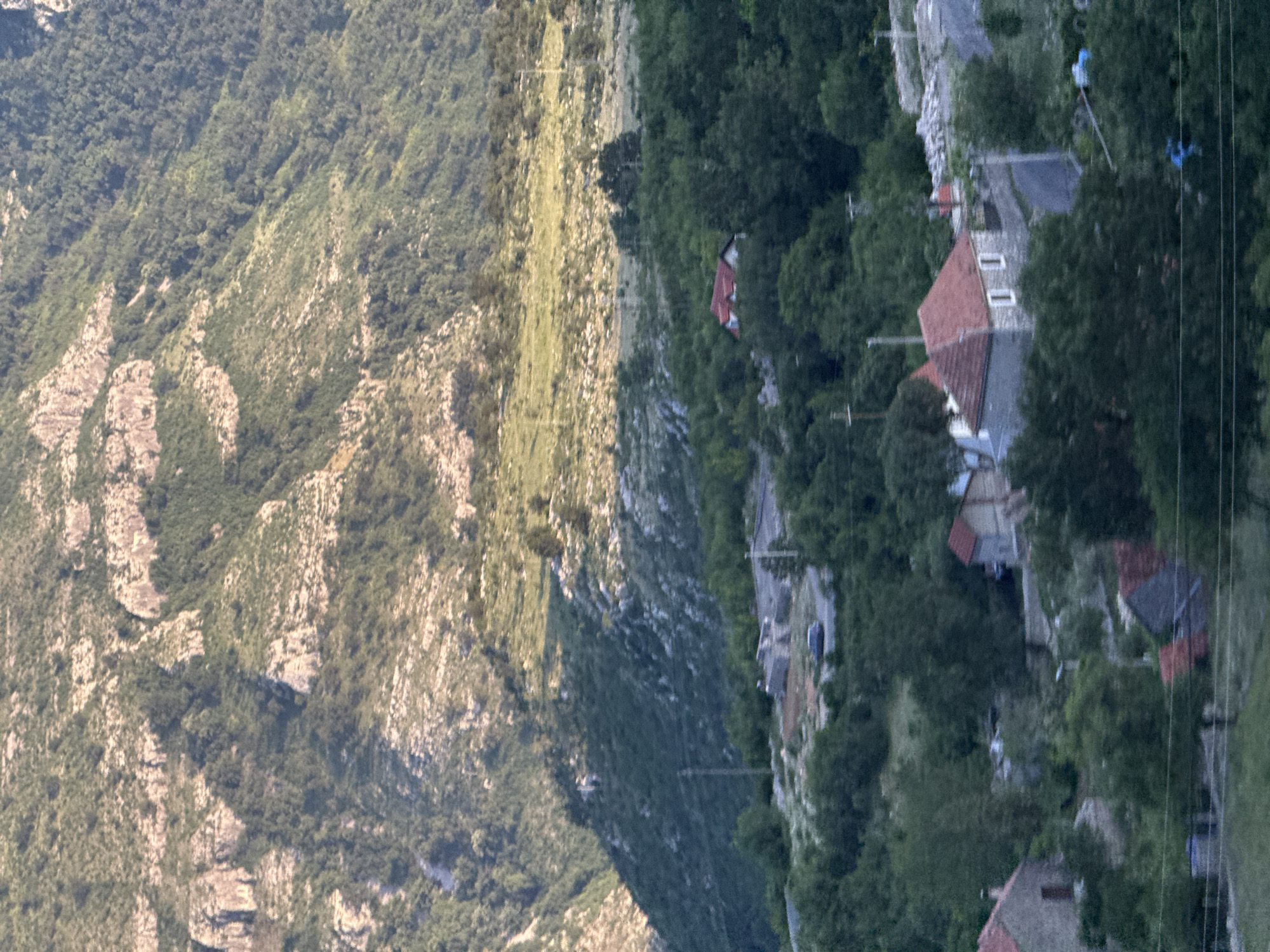 The image depicts the mountainous landscape of Klopot, Montenegro. On the left, a steep, rocky mountainside is visible, densely covered with vegetation. To the right, a small village is nestled among the trees, with several houses featuring distinct red-tiled roofs. A road winds through the village, and power lines are strung overhead. The lighting suggests it is daytime.